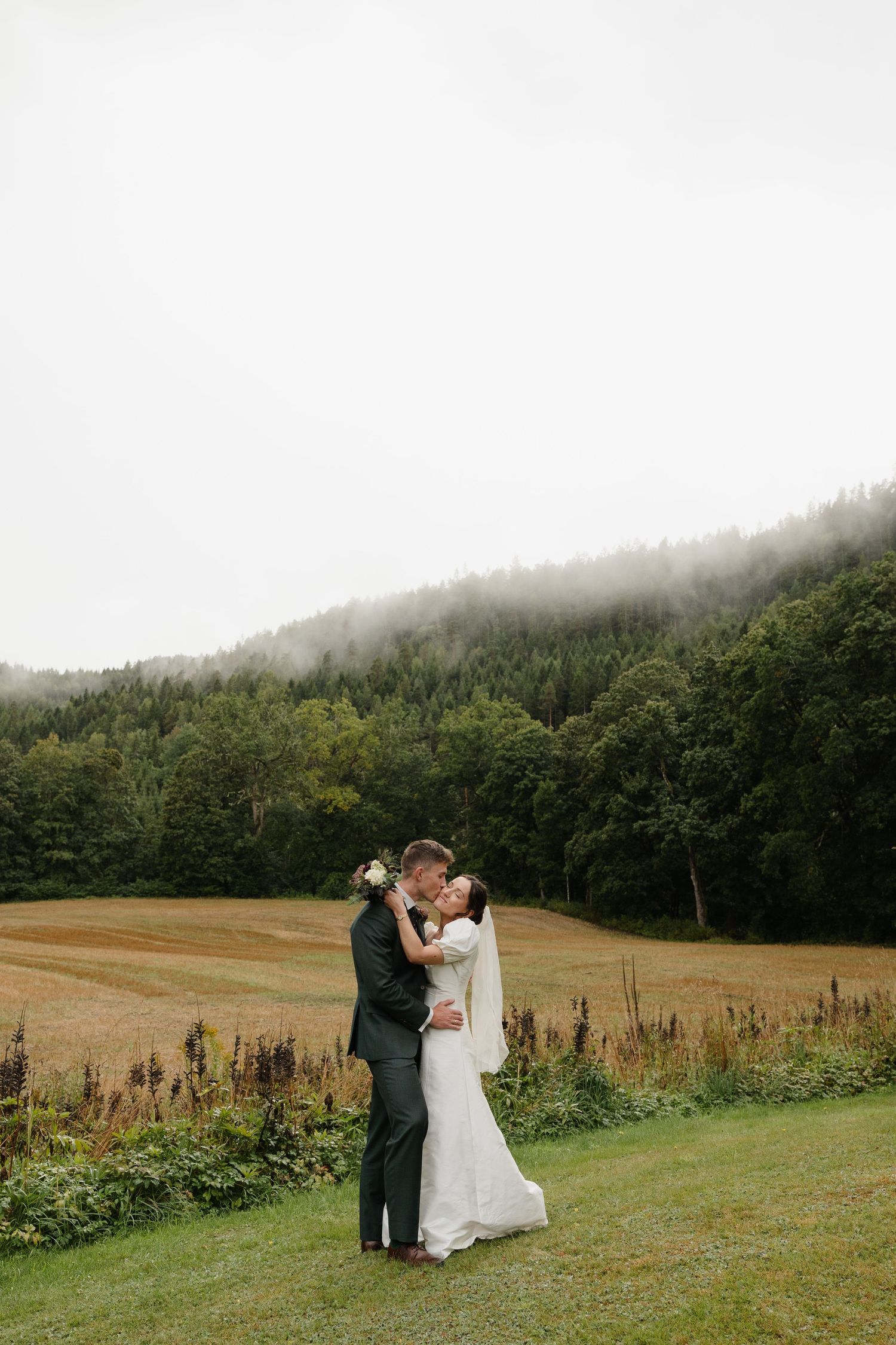 Bride and groom embracing in a misty field with foggy forested hills behind them.