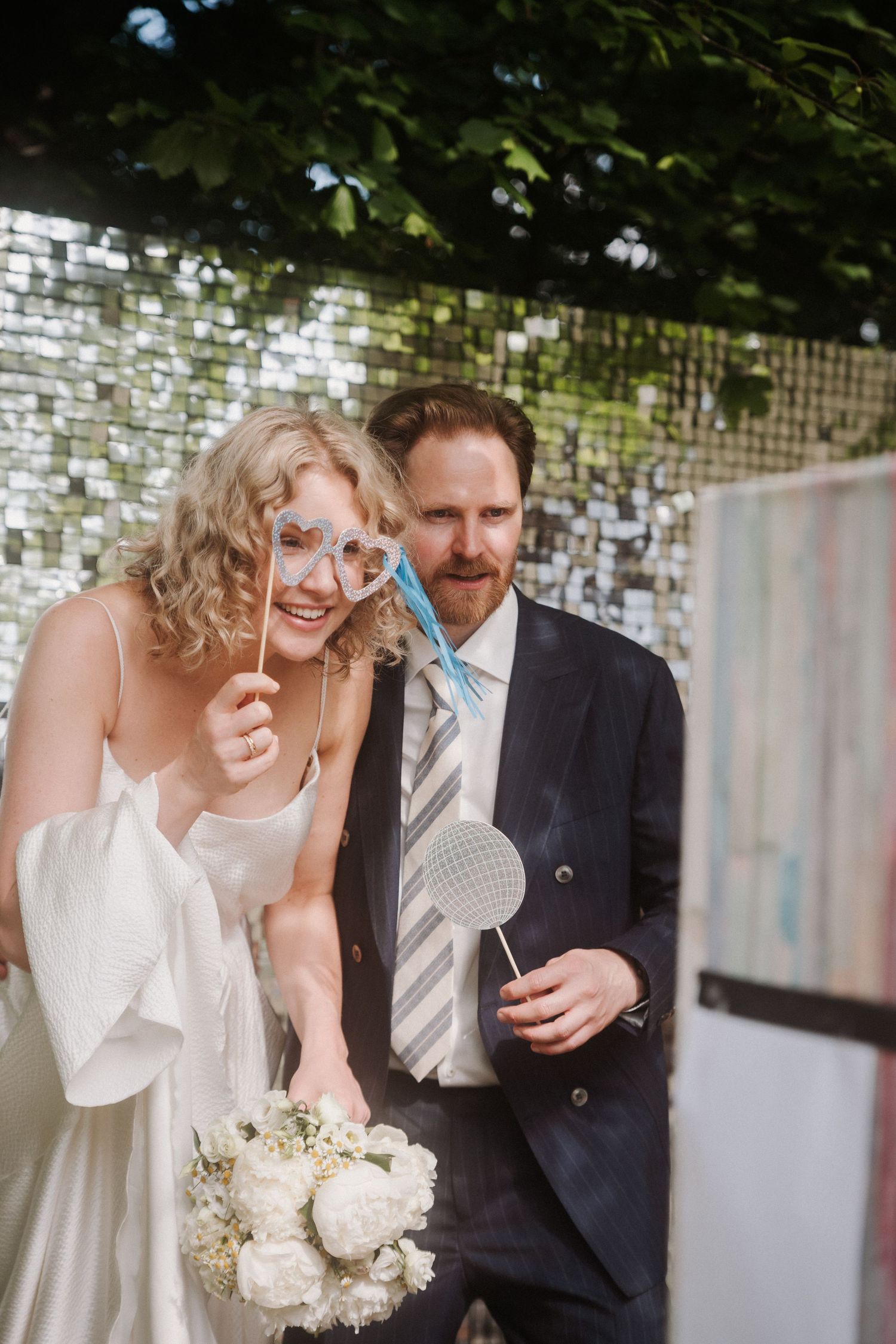 A bride and groom pose playfully with photo booth props in front of a shimmering sequin backdrop.