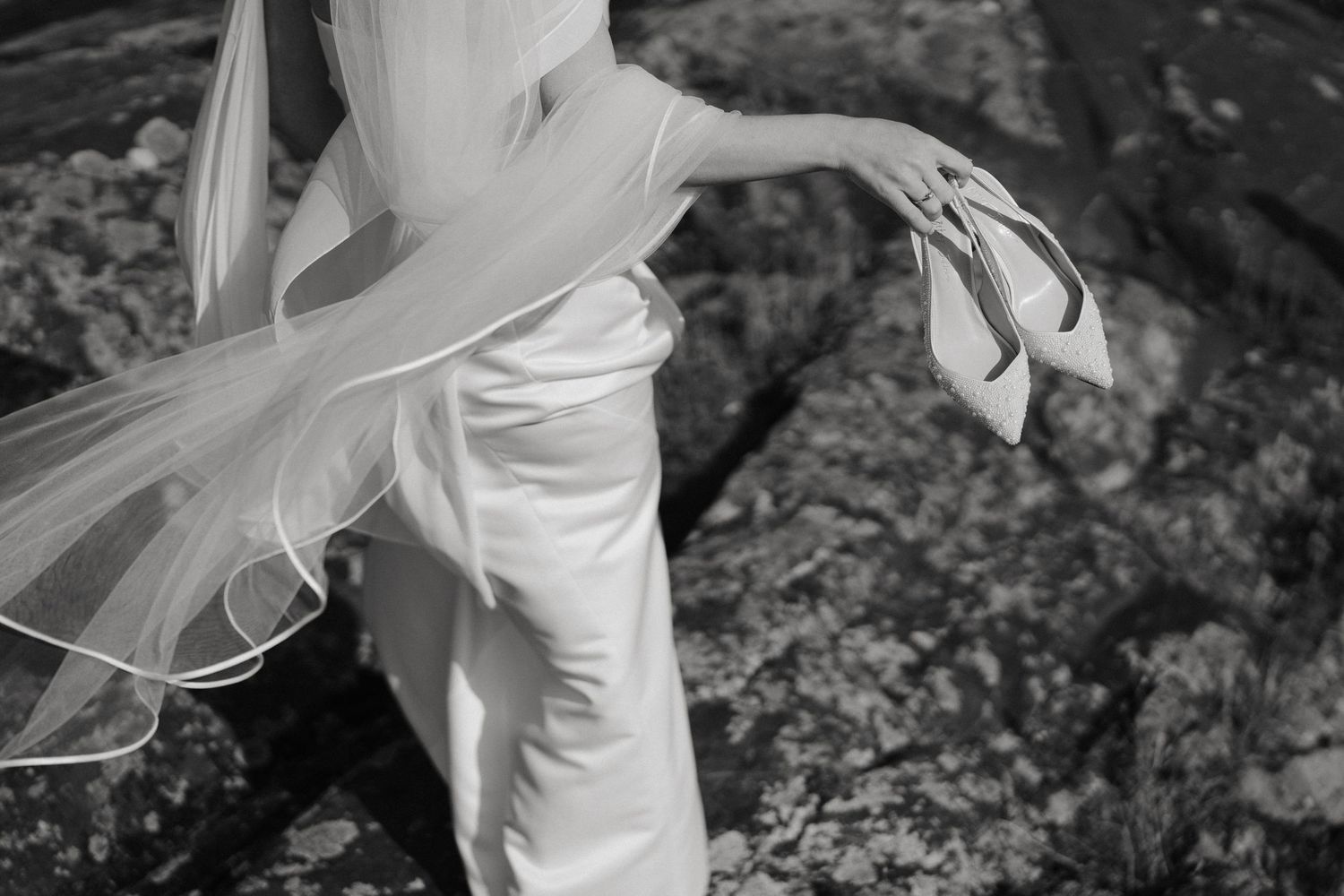 Black and white image of a bride in a white dress holding heels on rocky ground.