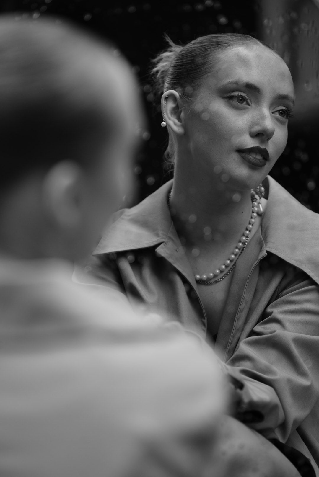 Black and white portrait of a woman wearing a pearl necklace and dark lipstick, gazing thoughtfully.