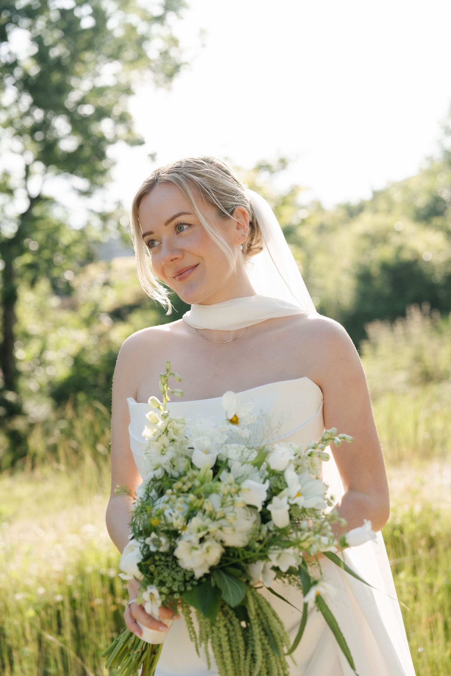 Bride in strapless white gown holding lush white floral bouquet outdoors in sunlit green field.