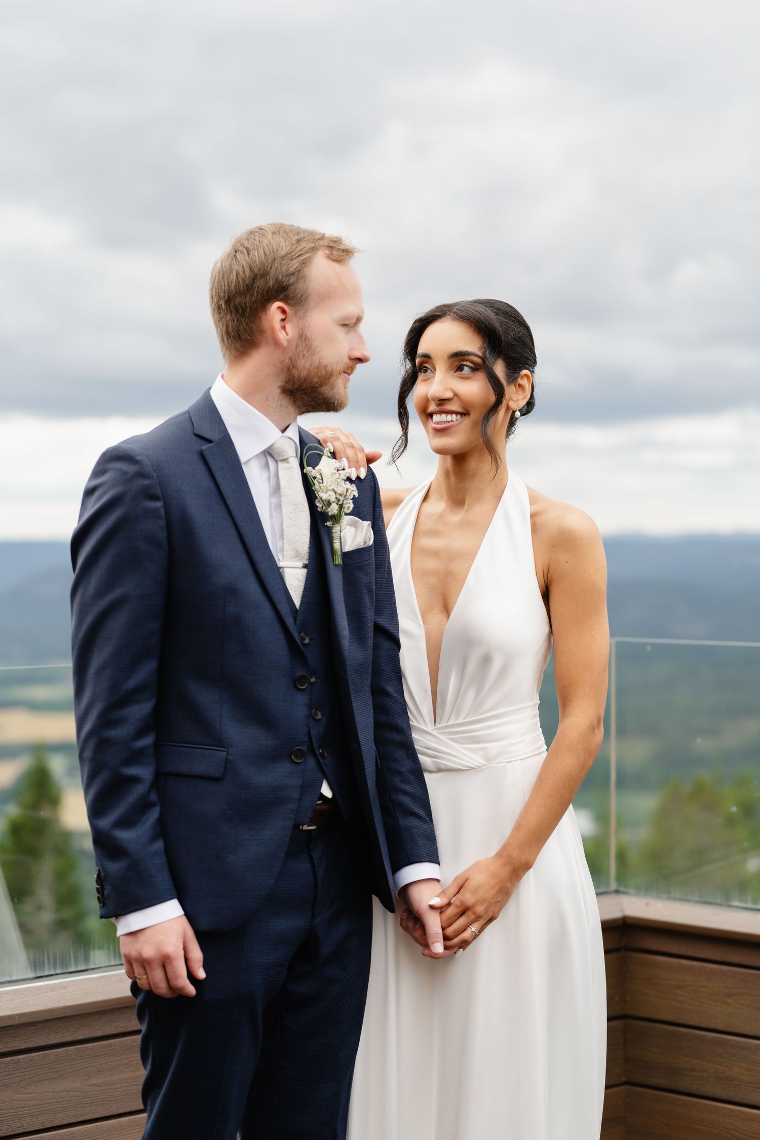Smiling bride in deep-V white gown holds hands with groom on elevated deck with mountain views.