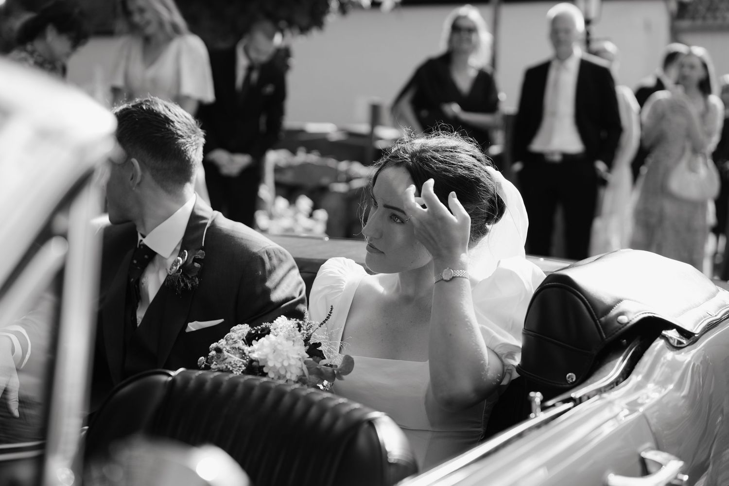 Bride adjusting her veil in a vintage car while groom sits beside her at a wedding.