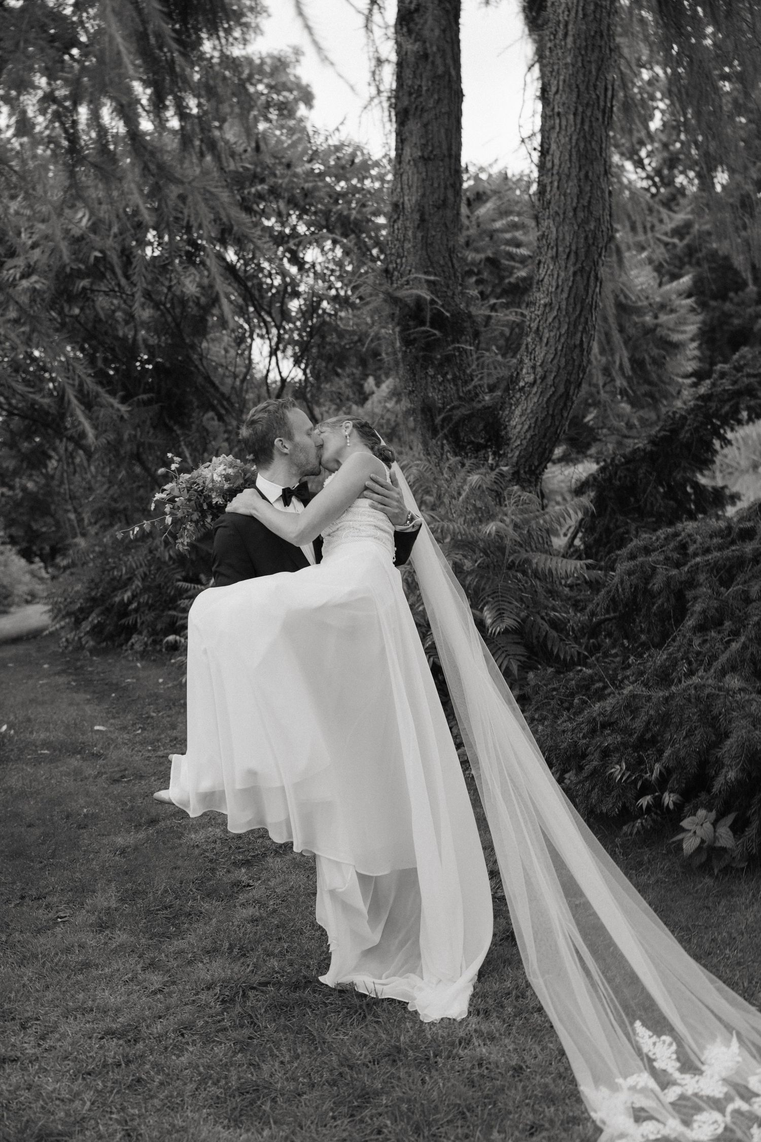 Black and white photo of groom lifting bride for a kiss beneath tall trees in a garden.