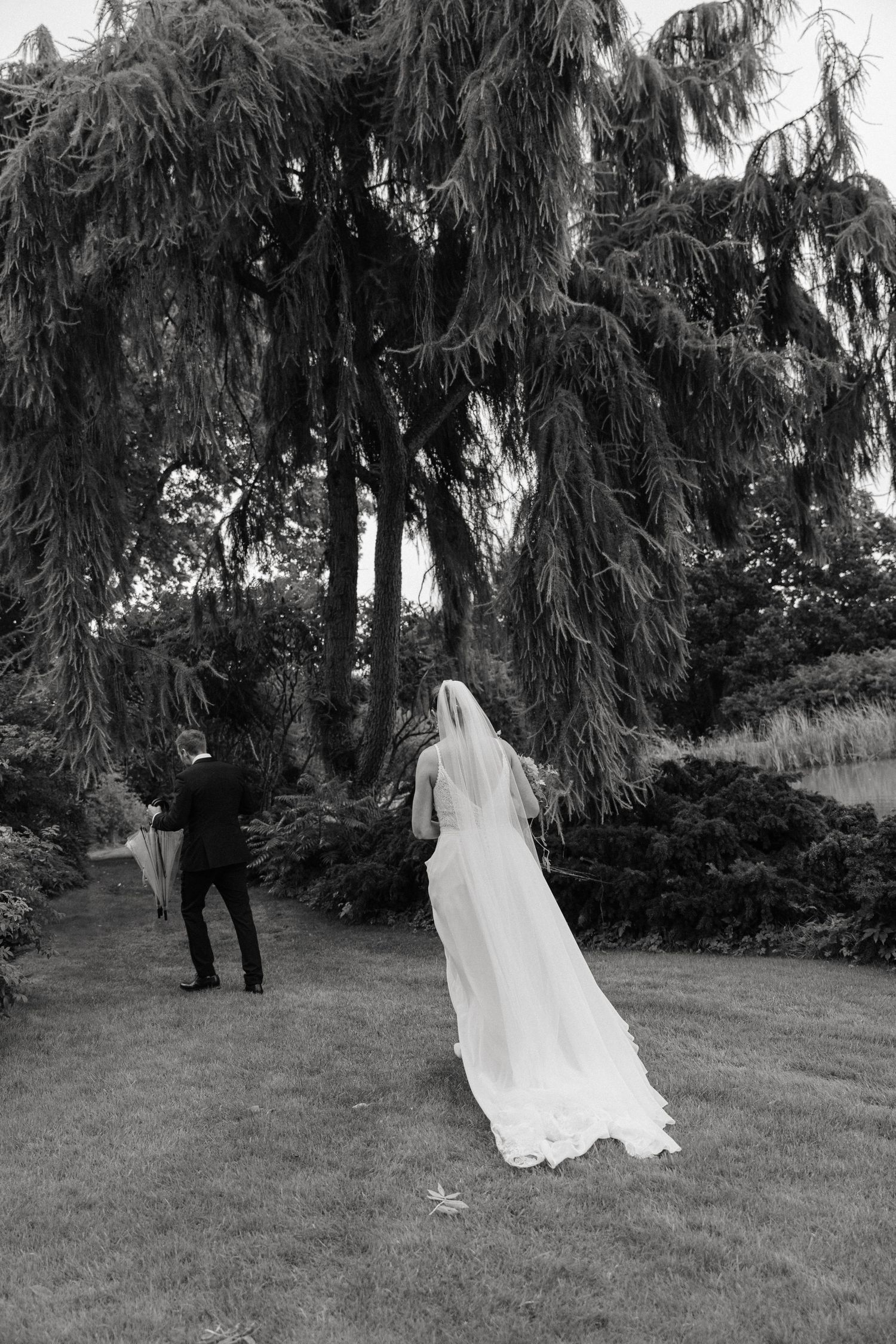 Black and white photo of bride and groom walking toward weeping willow tree in lush garden.