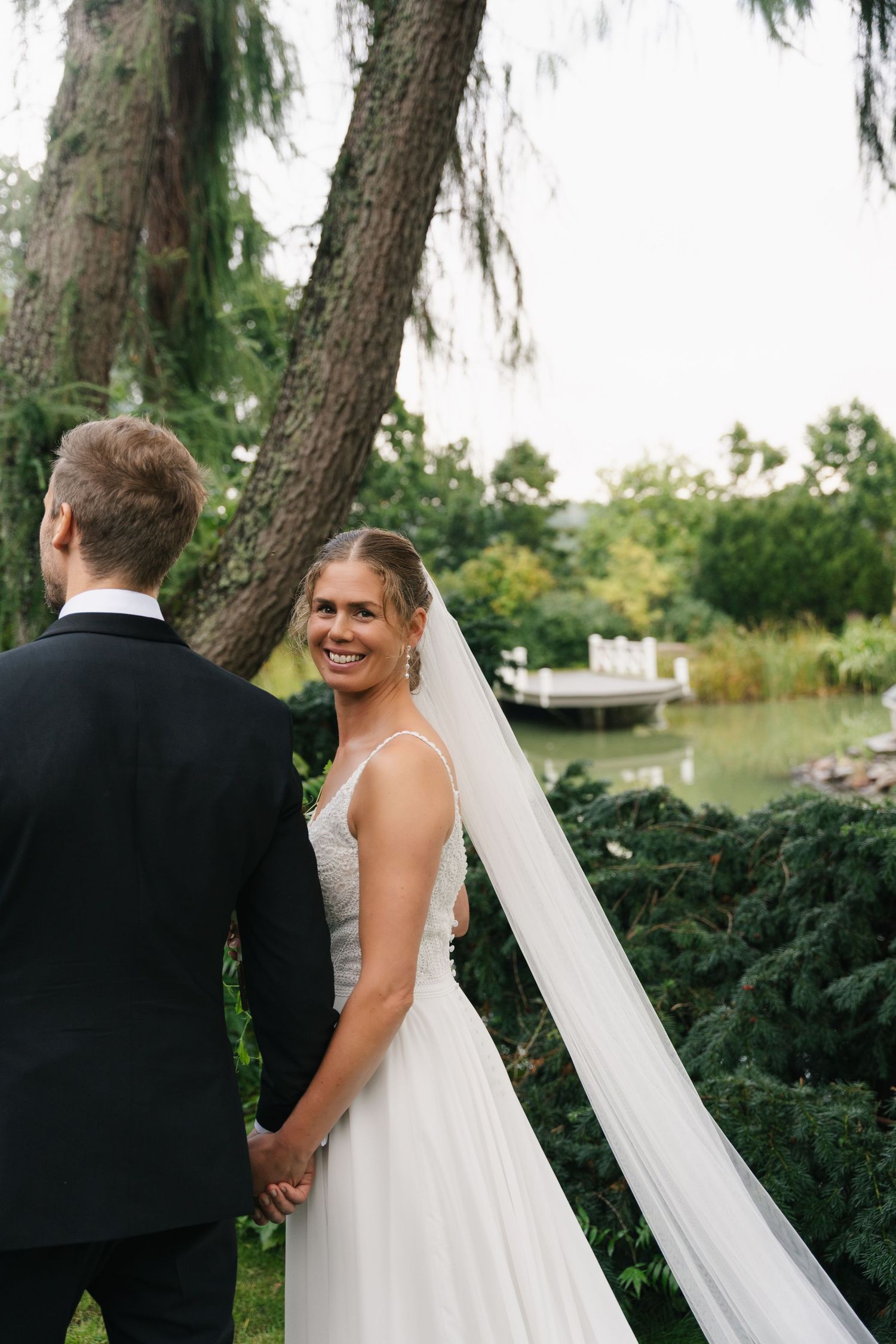 Smiling bride in white gown and veil holds grooms hand while looking back in a garden setting.