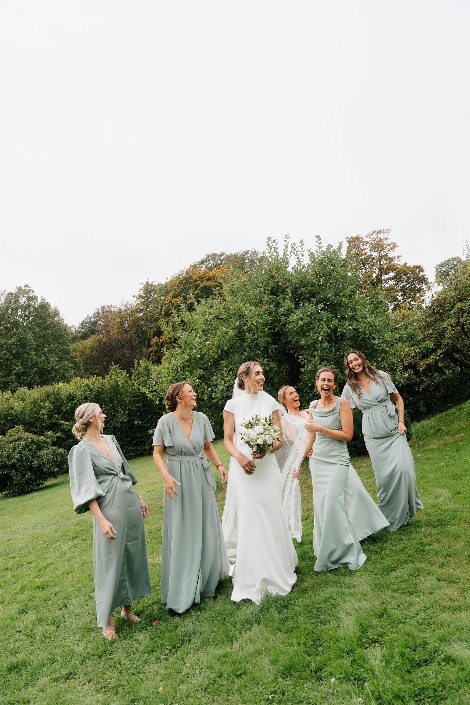 Bride in white gown walks outdoors with five bridesmaids in sage green dresses on a lawn.