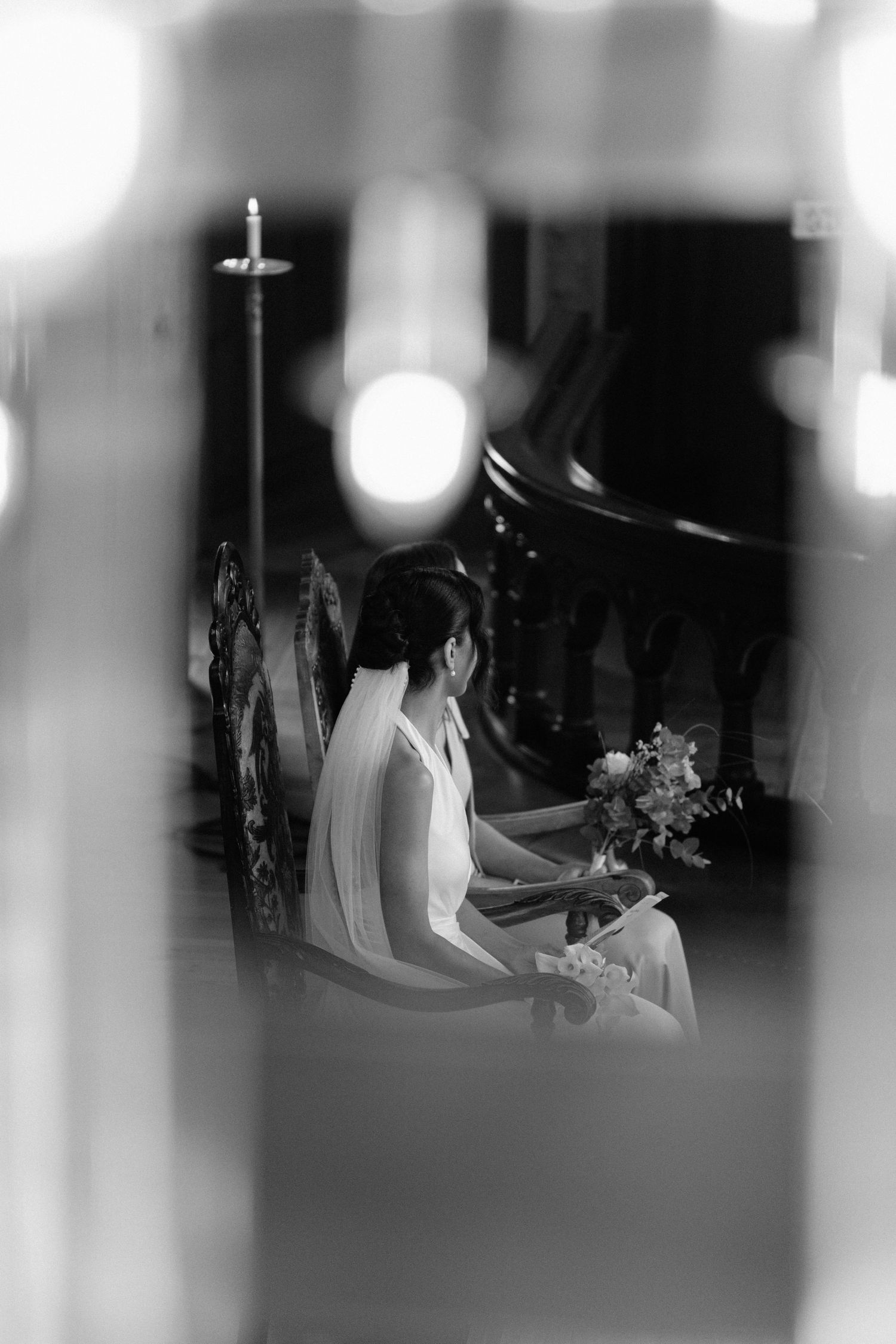 Black and white artistic shot of veiled bride seated in church holding a floral bouquet.