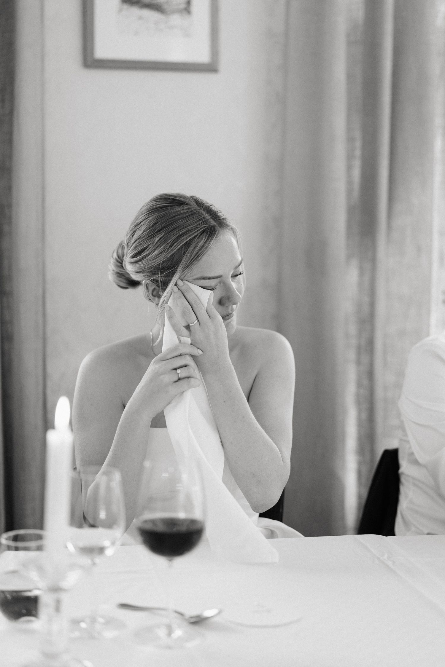 Black and white photo of an emotional bride dabbing tears with a napkin during wedding reception speeches.