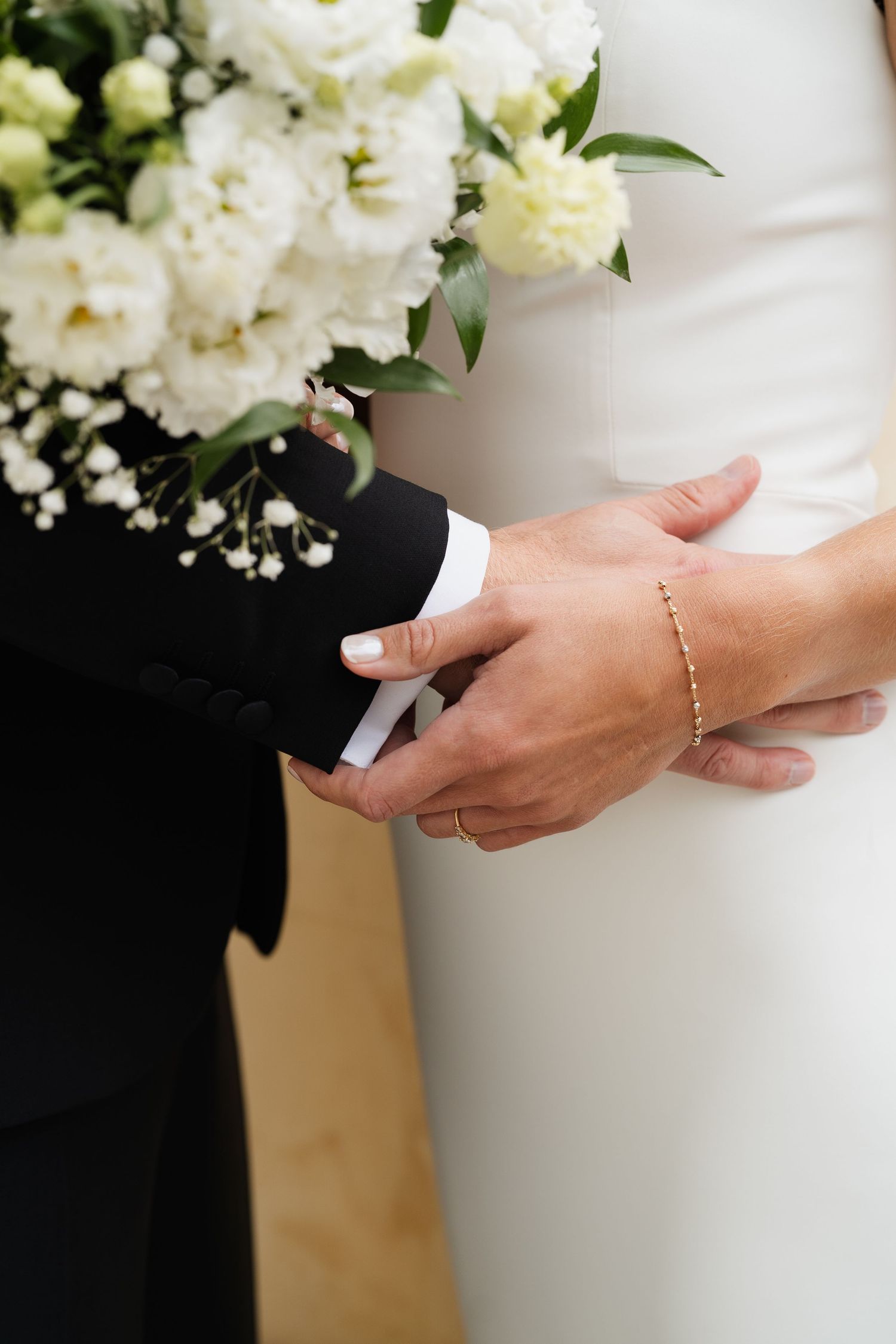 Close-up of a bride and groom holding hands, showcasing a gold bracelet and white floral bouquet.