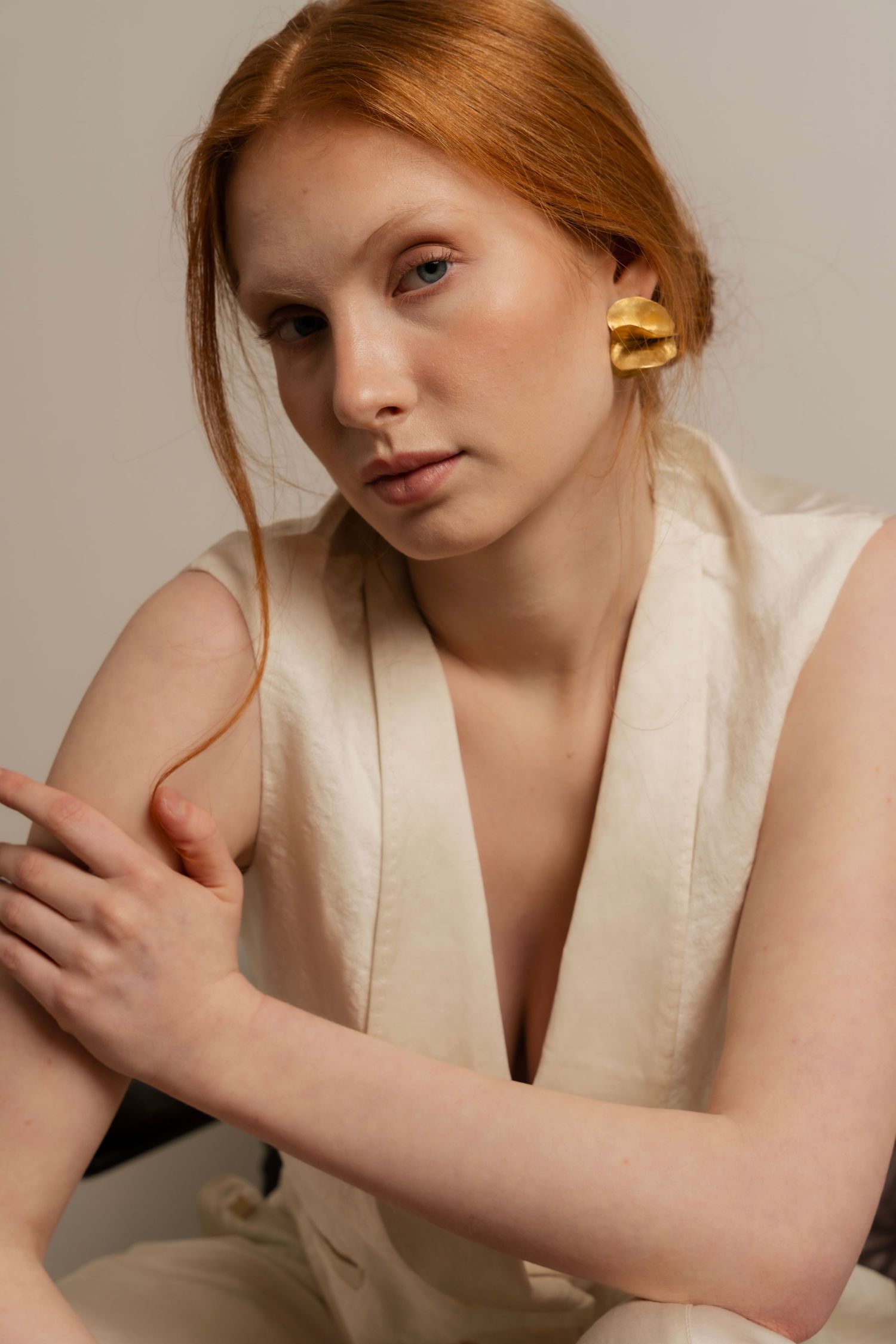 Red-haired woman wearing a gold earring and cream linen vest, posing gracefully against a neutral background.