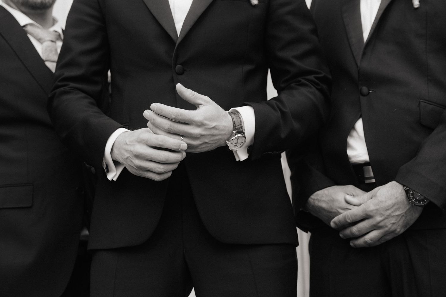 Three groomsmen in black suits stand together, showing clasped hands and watches in black and white.