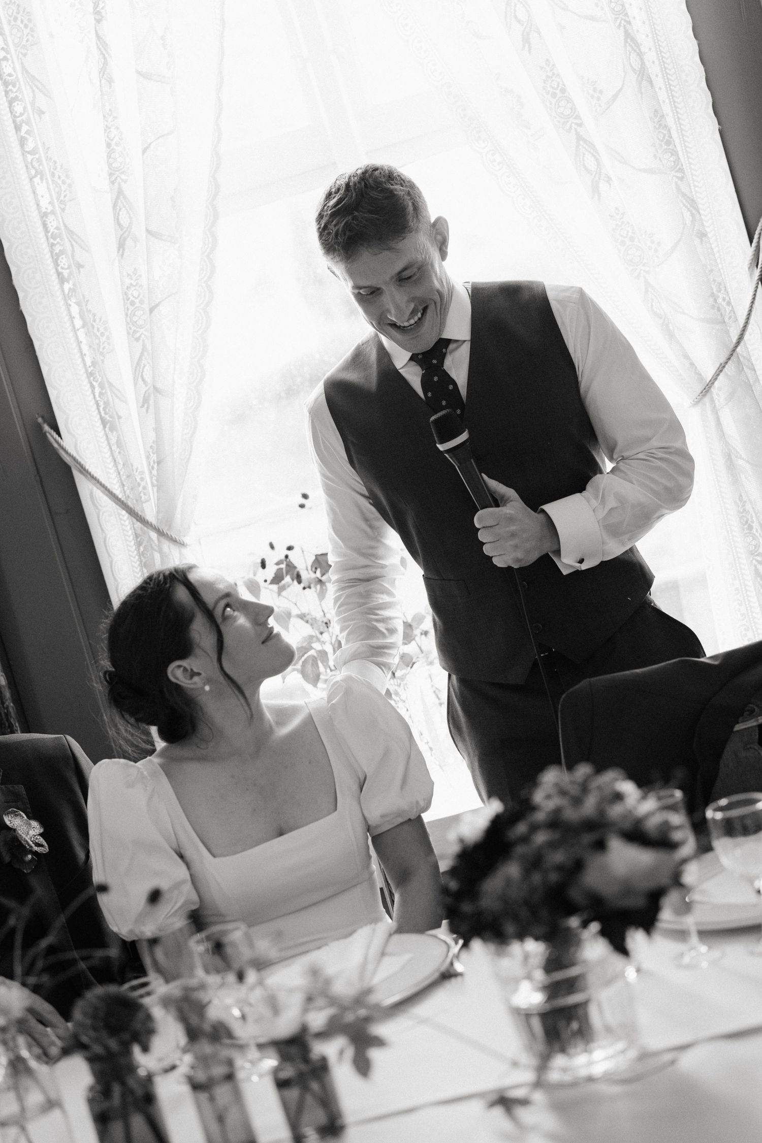 Black and white photo of smiling groom giving speech while bride looks up at him adoringly.