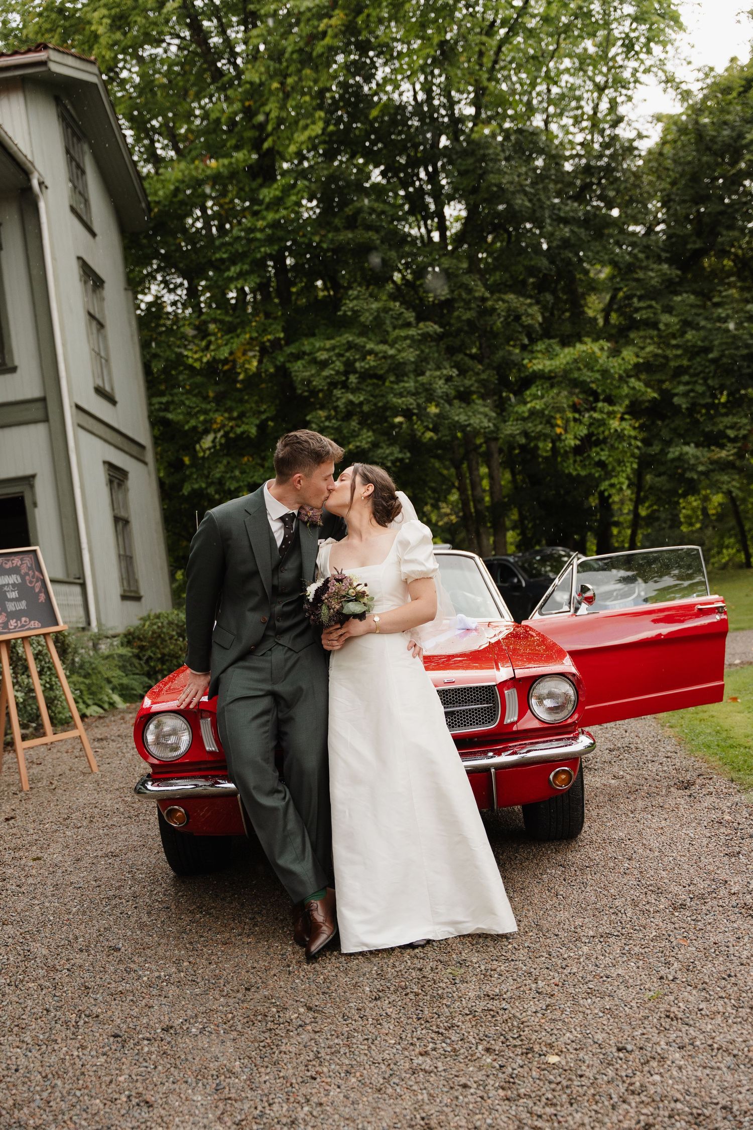 Bride and groom kissing beside a classic red Ford Mustang convertible outdoors.