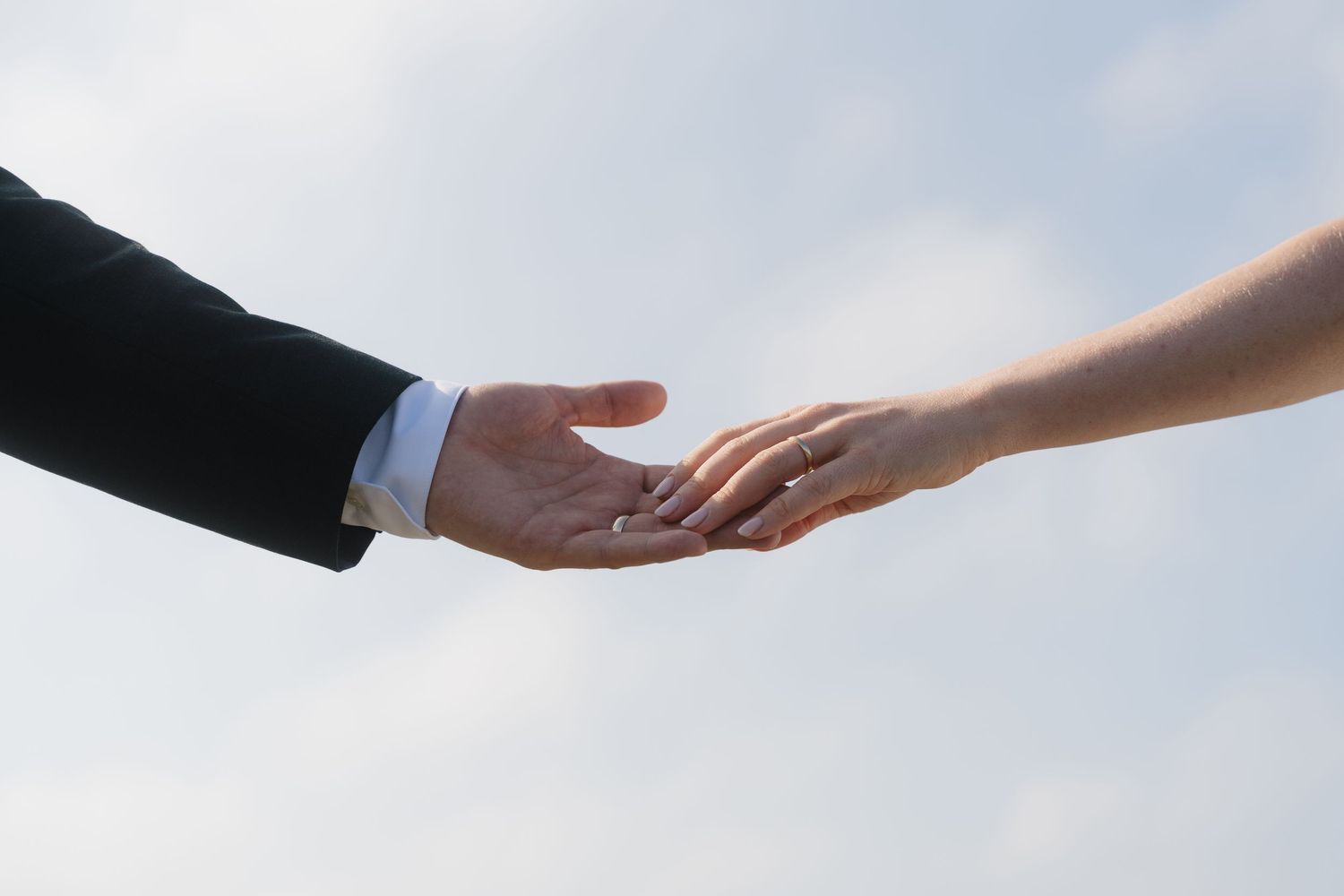 Close-up of a bride and groom reaching for each other's hands against a soft blue sky on their wedding day.