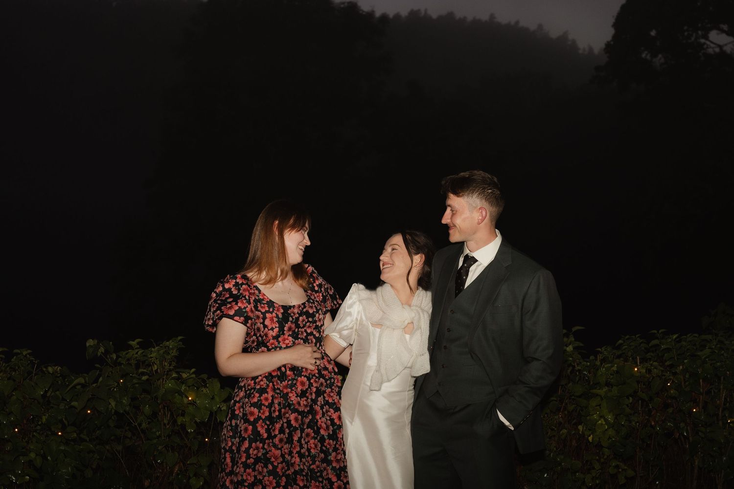 Bride in white dress with two guests smiling outdoors at night during wedding celebration.