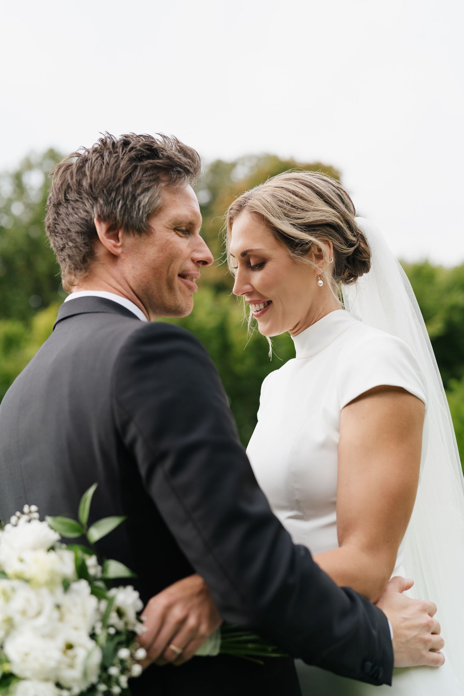 Smiling bride and groom embrace outdoors, with lush greenery in the background on their wedding day.