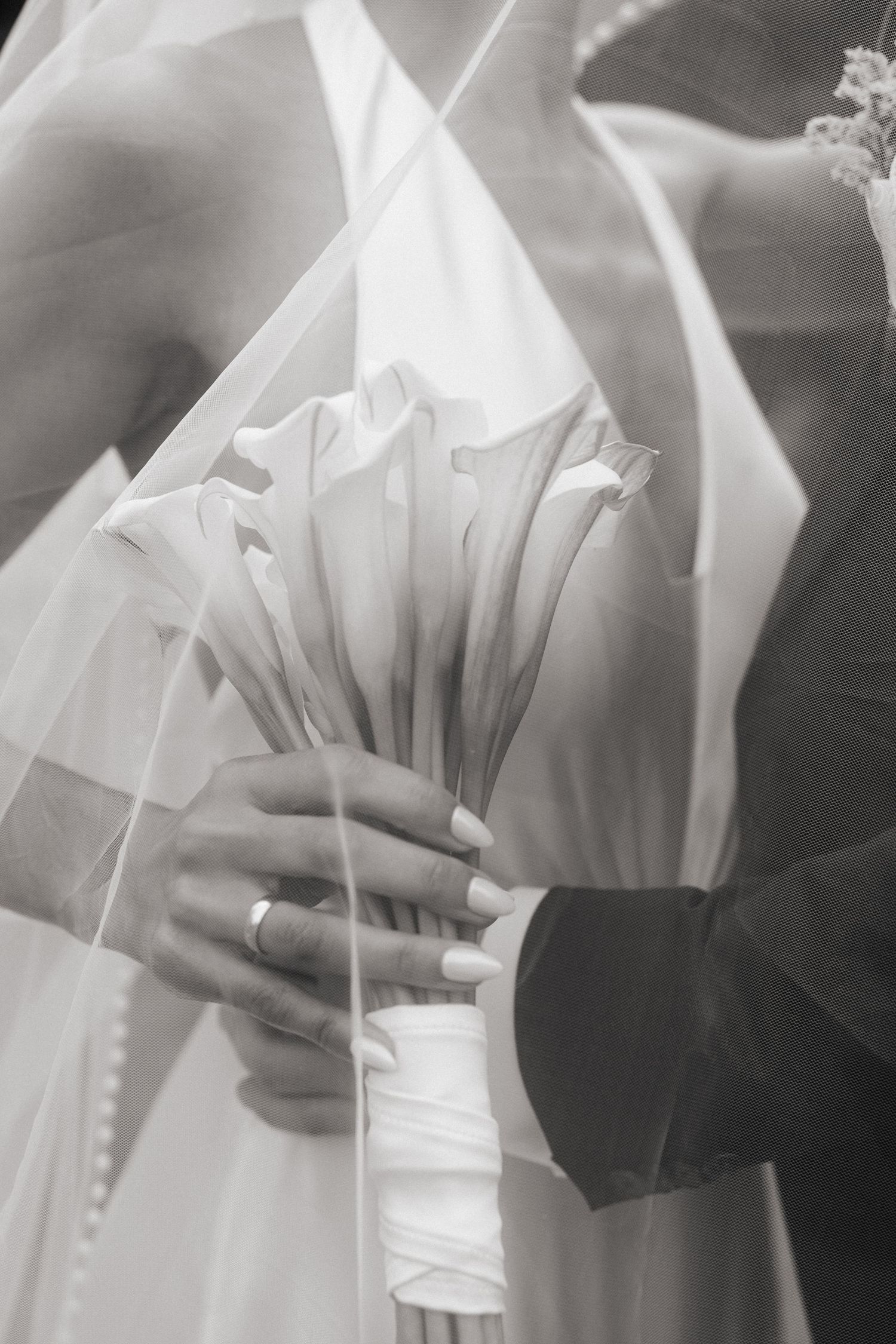 Black and white wedding photo of bride and groom holding calla lily bouquet, showing wedding rings and bridal veil.