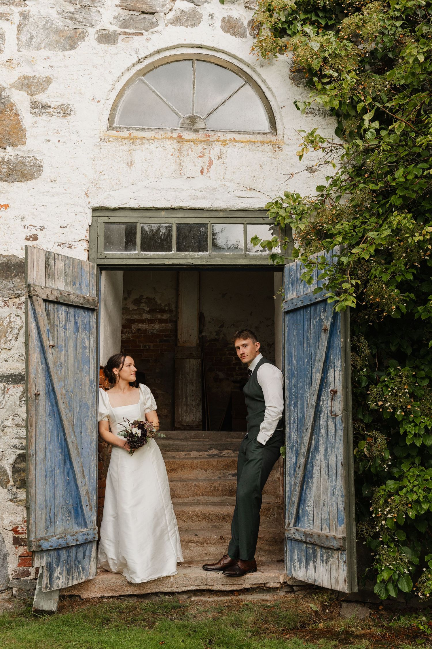 Bride and groom stand in rustic stone doorway with blue wooden doors and greenery.