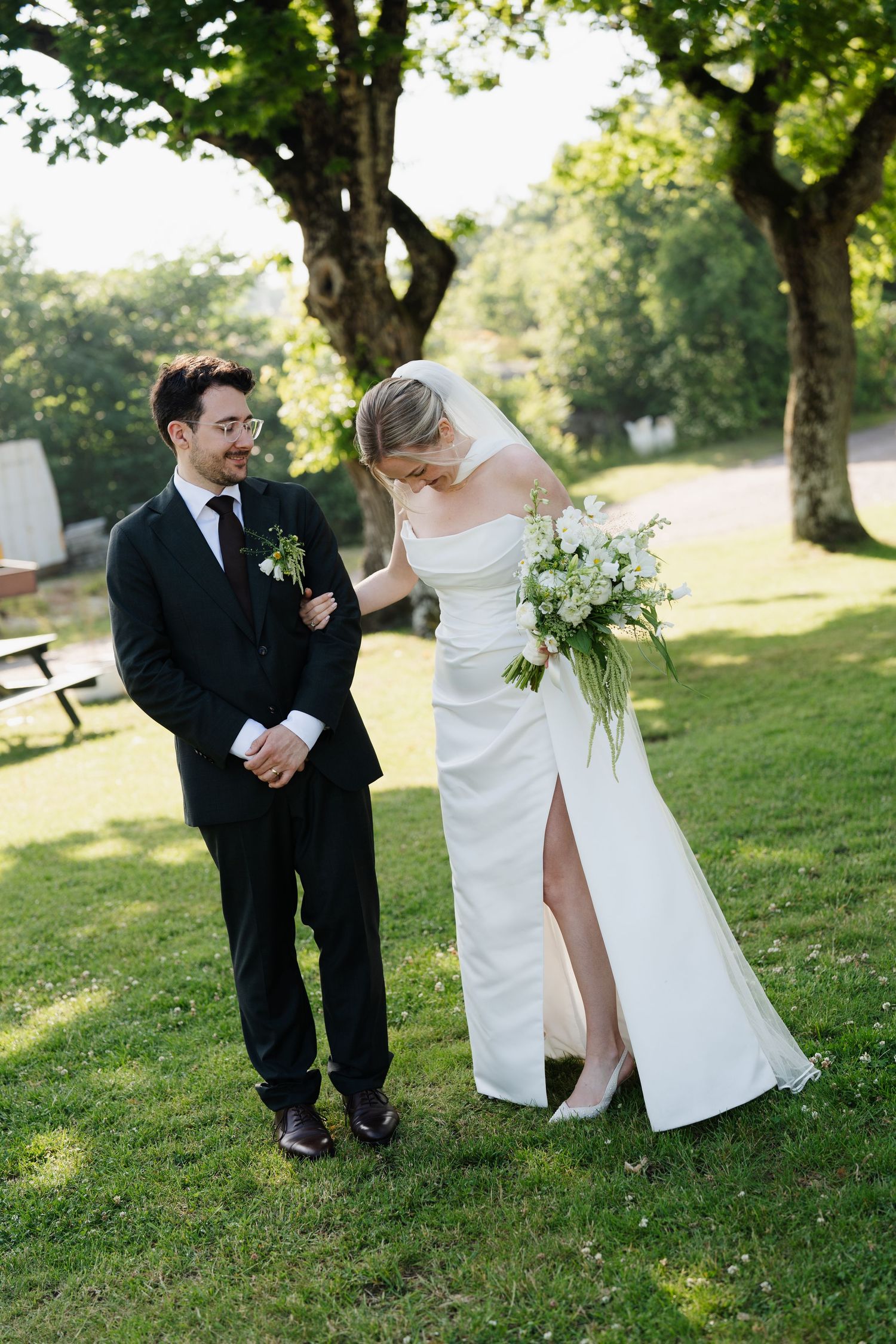 Groom in black suit smiling at bride in white gown with slit in sunny garden setting.