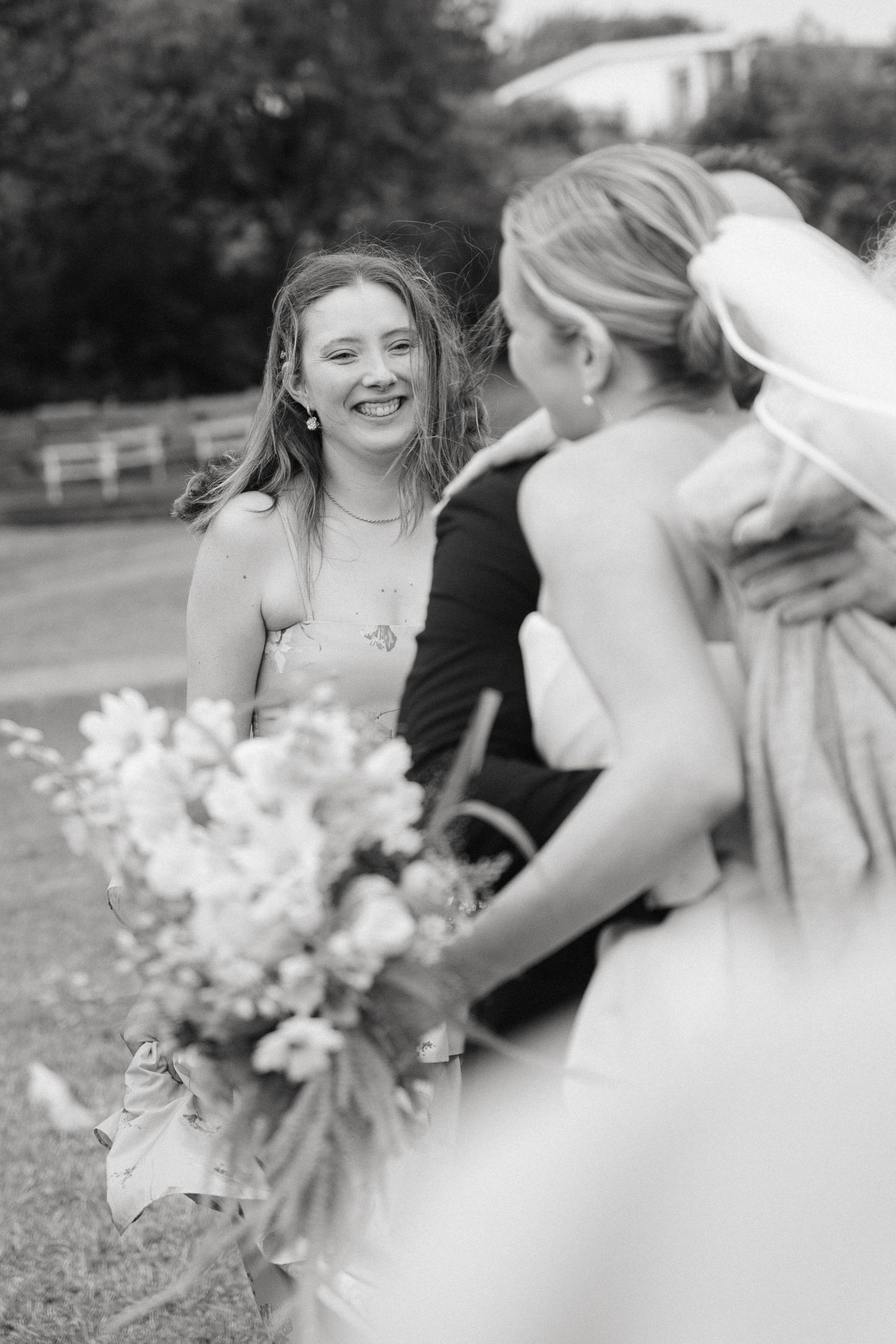 Black and white wedding photo of smiling bridesmaid holding floral bouquet while chatting with bride outdoors.