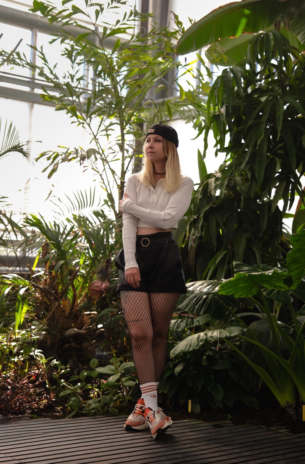 Young woman in white top, black skirt, fishnet tights, and cap poses in a lush tropical greenhouse.