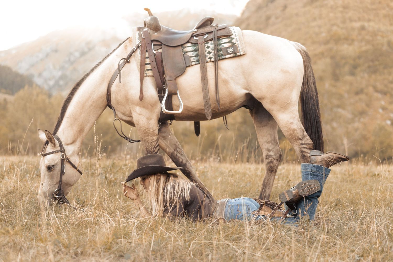 Woman in cowboy hat kneeling beneath a saddled palomino horse grazing in a golden meadow with mountains behind.