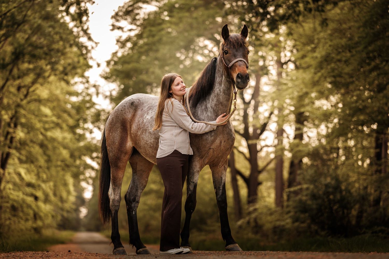 Woman in white blouse stands beside a brown roan horse on a tree-lined path in autumn.