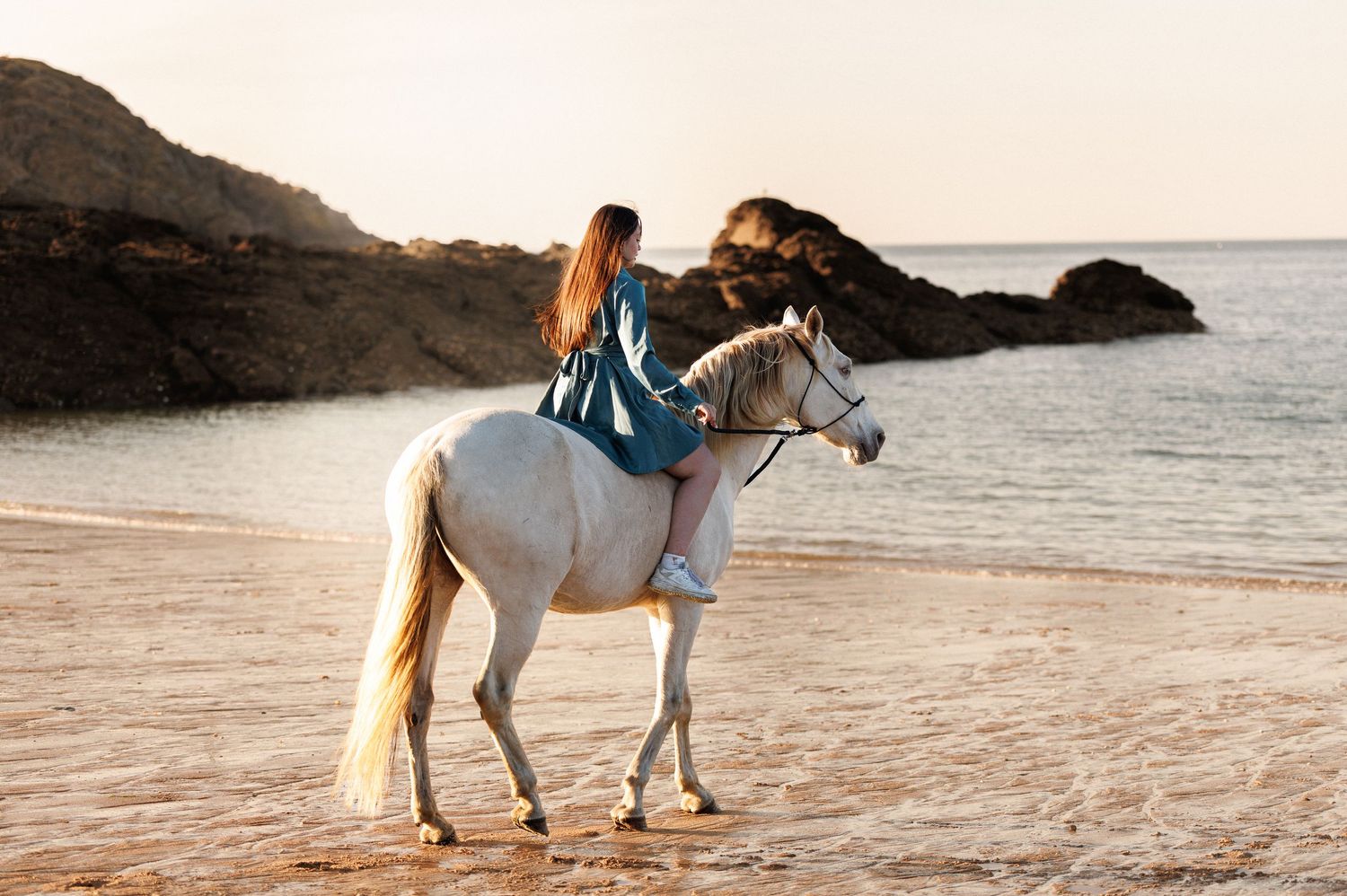 Woman riding a white horse on a sandy beach at sunset with rocky cliffs and calm ocean waves in the background.