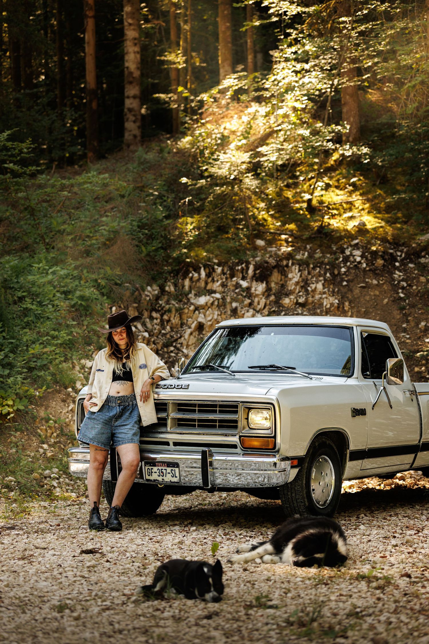 Woman in cowboy hat leaning on vintage Dodge pickup truck in forest with two dogs on gravel path.