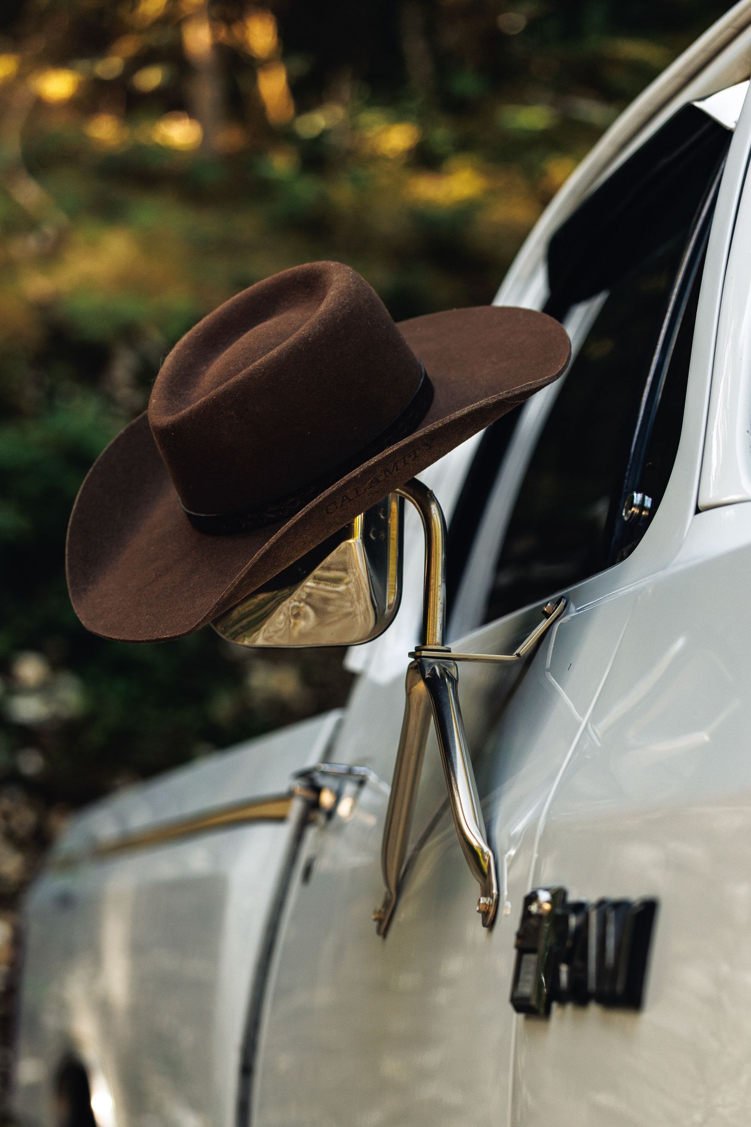 Brown cowboy hat resting on a vintage car side mirror, with blurred autumn forest background.