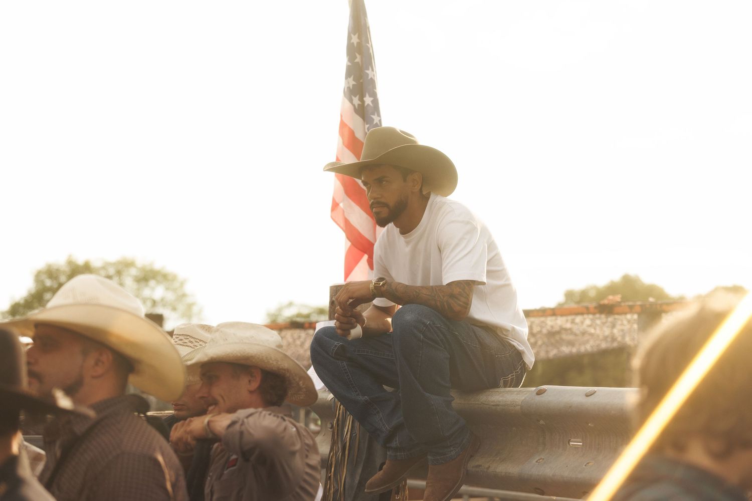 Man in white shirt and cowboy hat sits on truck with American flag at outdoor country gathering in golden hour light.