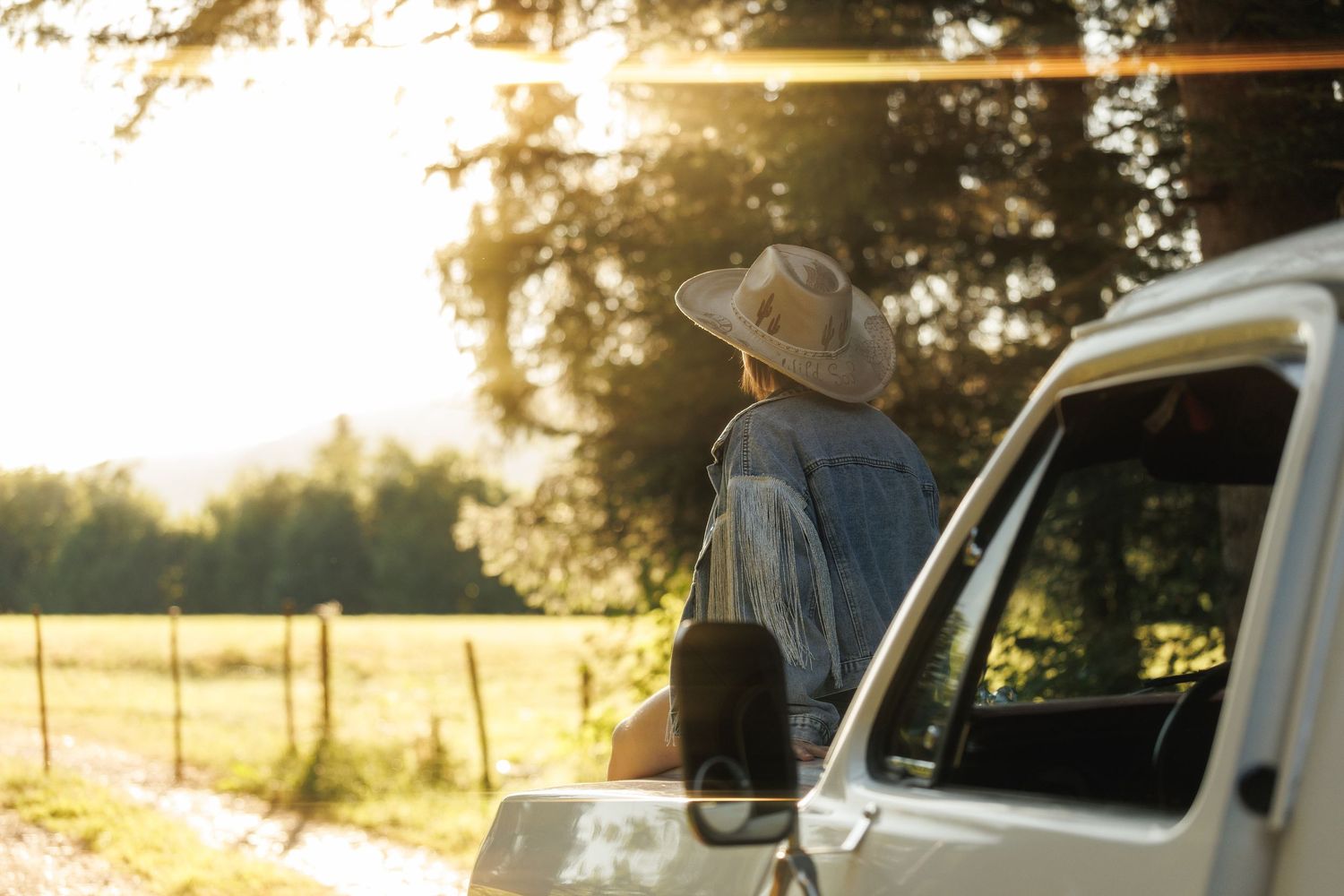 Person in a wide-brim hat leaning on a white truck, gazing at a sunlit rural field at golden hour.