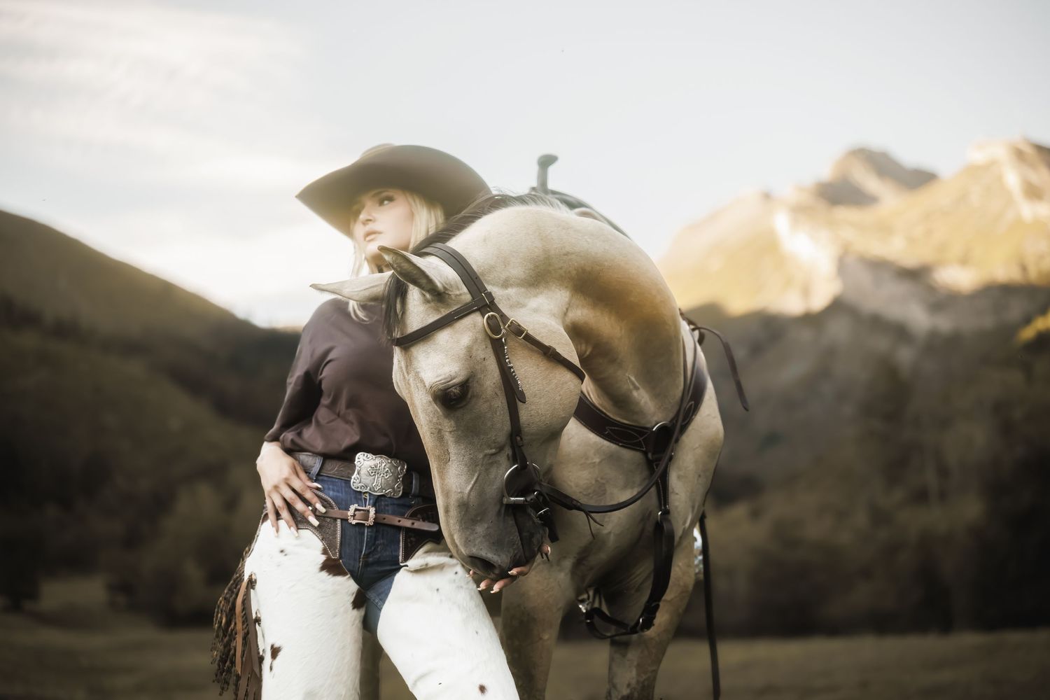 A cowgirl in a hat and white pants stands beside a gray horse with mountain scenery in the background.