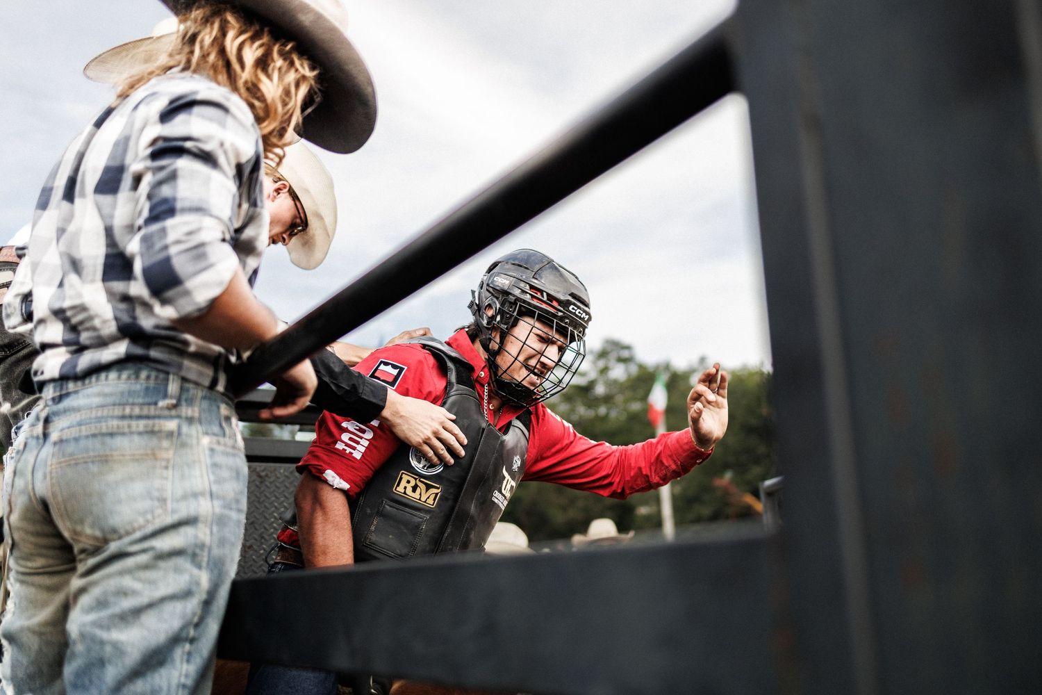 Bull rider in red vest and helmet prepares in chute assisted by cowboy in plaid shirt at rodeo event.