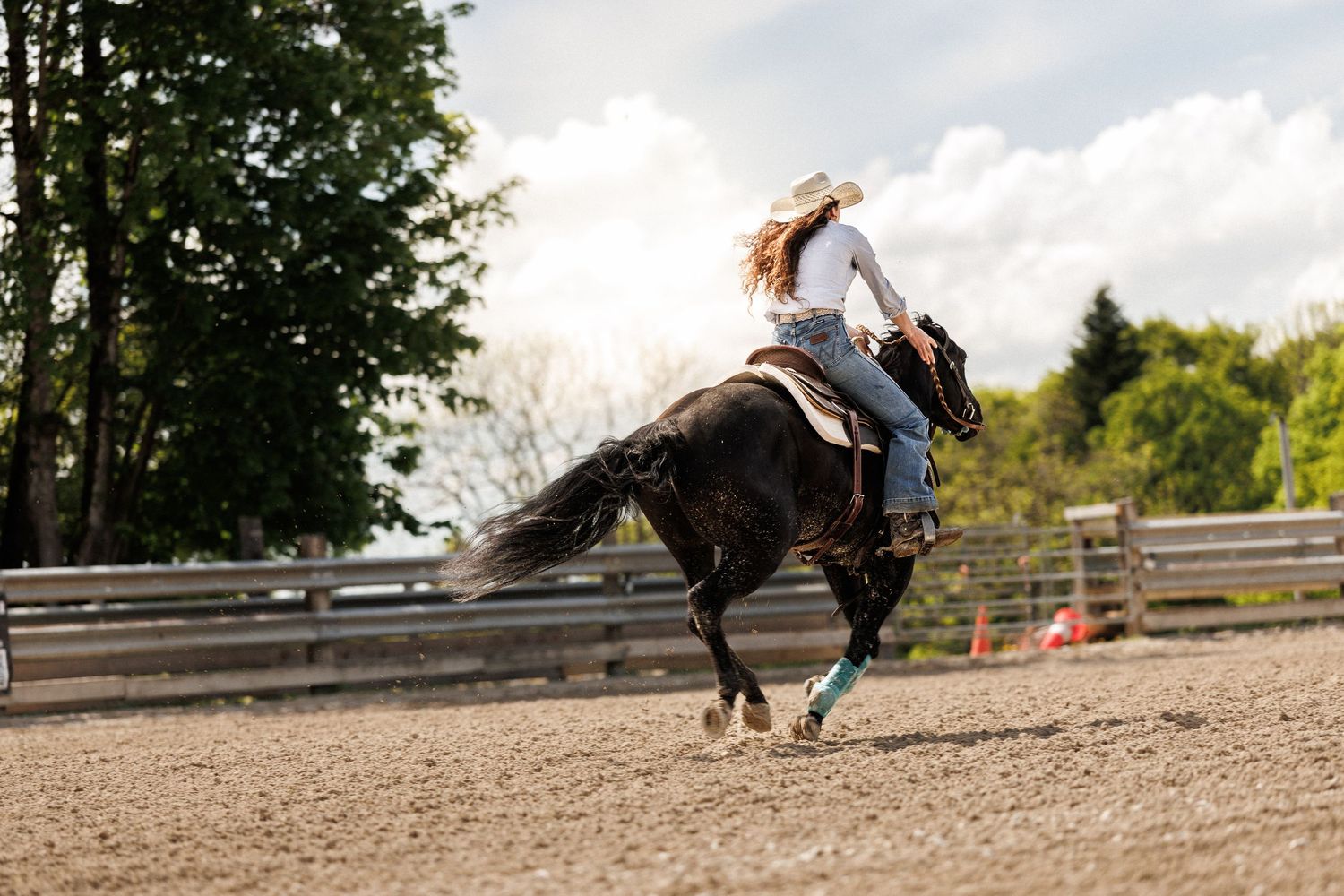 Woman in cowboy hat riding a dark horse in a sandy arena during a western equestrian event.