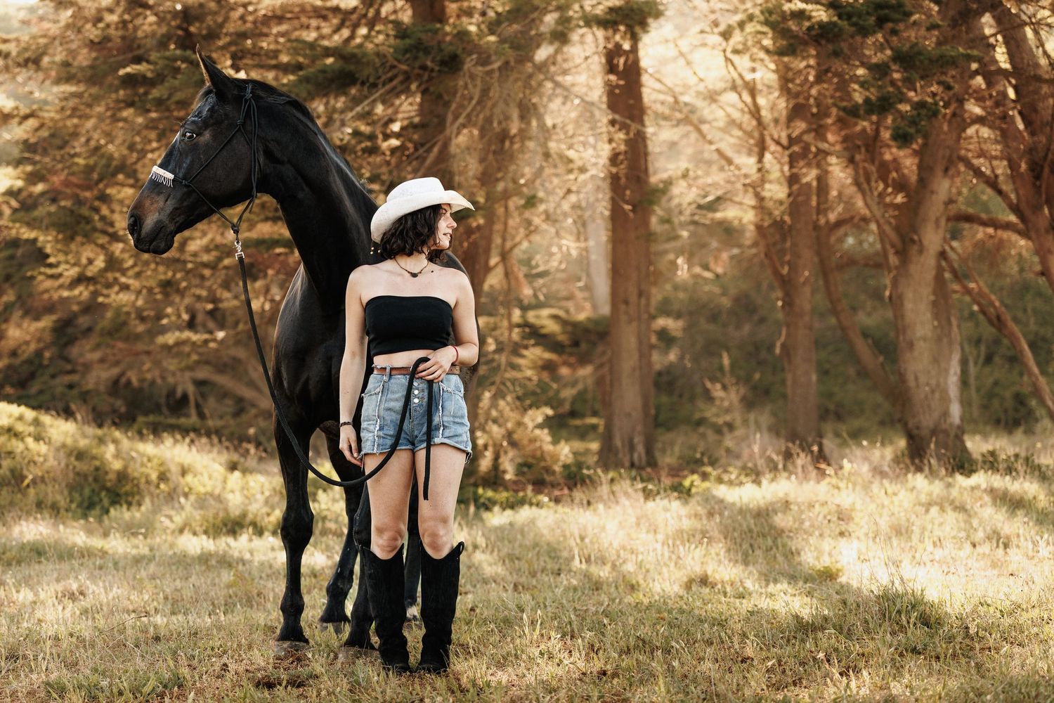 Young woman in cowboy hat, black tube top, denim shorts, and boots standing beside a black horse in a forest.