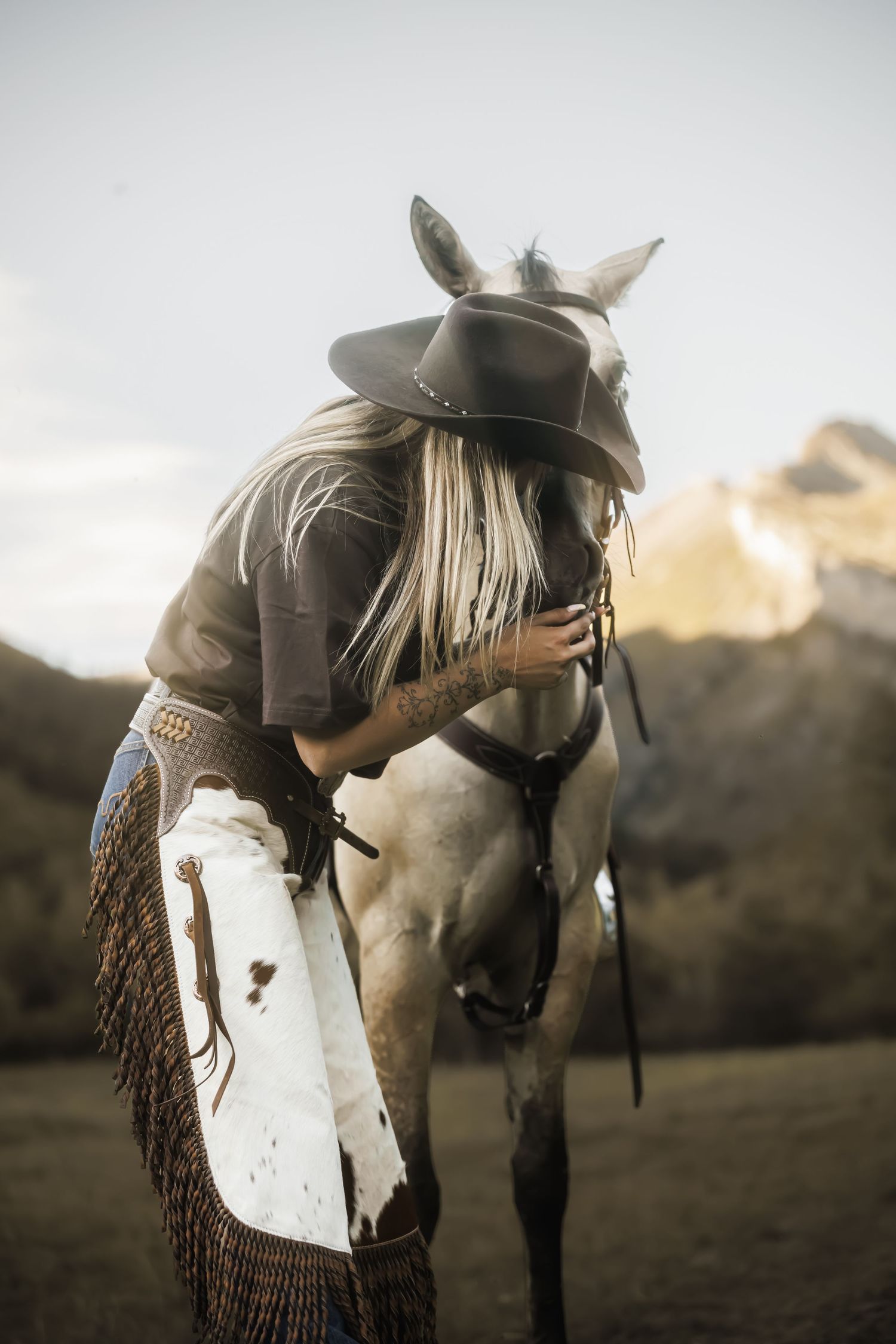 A cowgirl in a wide-brim hat and chaps embraces her gray horse in a scenic mountain landscape.