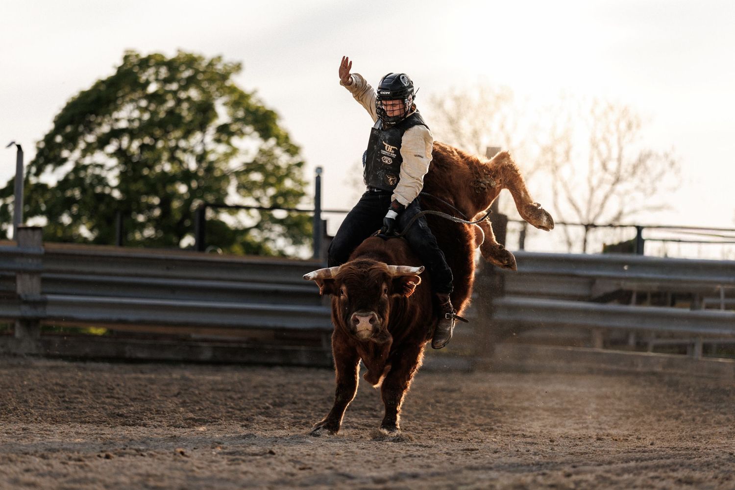 A rodeo rider in protective gear attempts to stay on a bucking brown bull in a dusty arena.