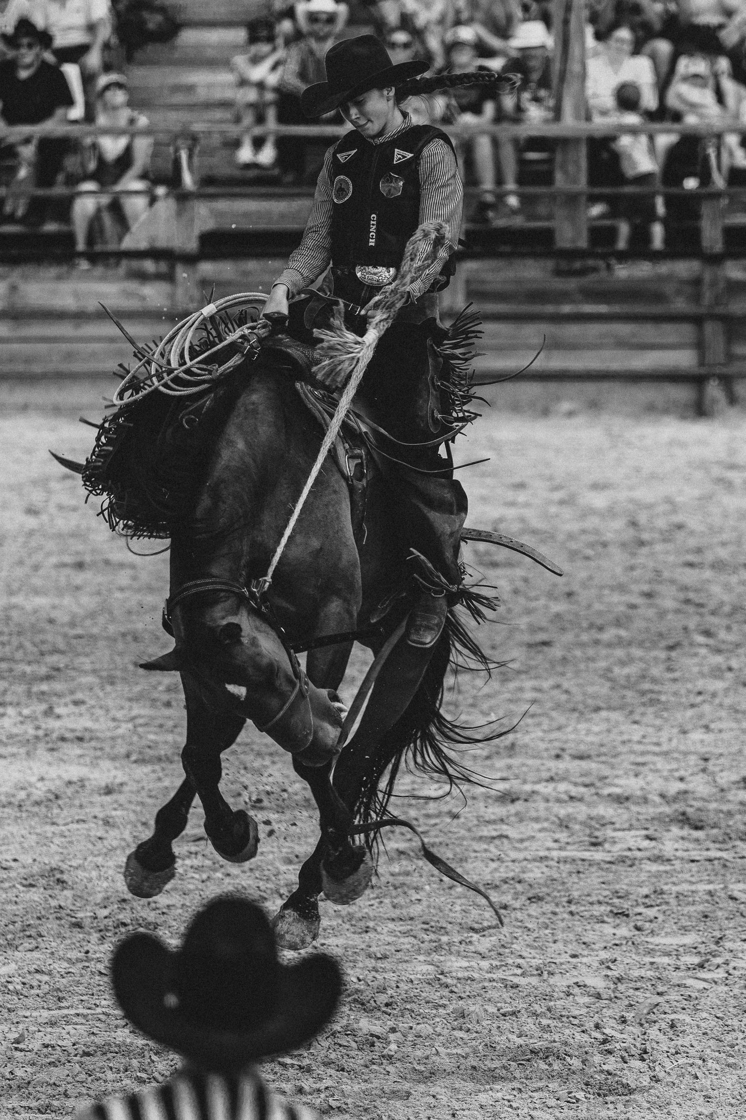Black and white photo of a cowgirl riding a horse at a rodeo, holding a lasso with crowd watching in background.