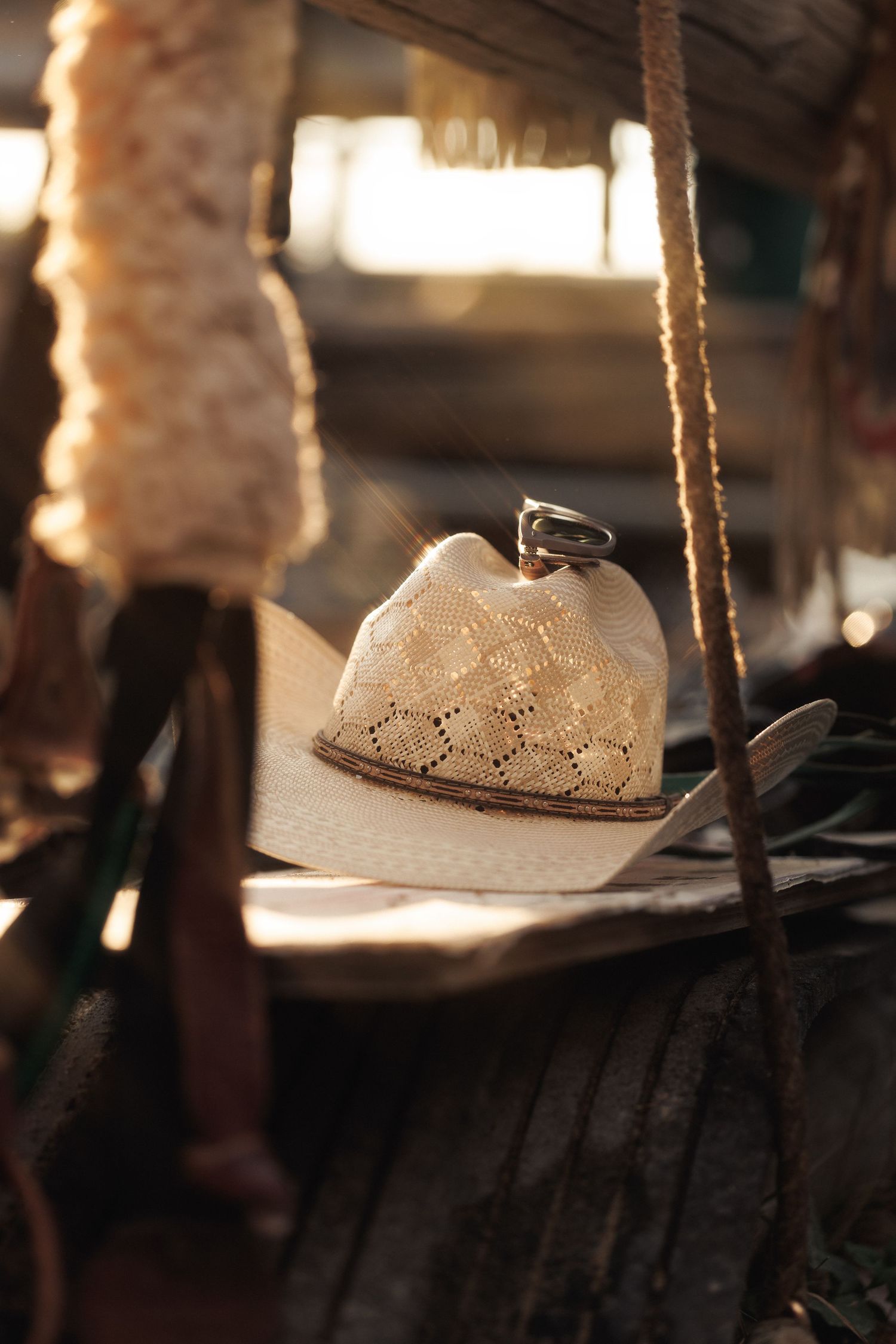 A straw cowboy hat with a silver ring rests on a wooden fence in warm golden sunlight.