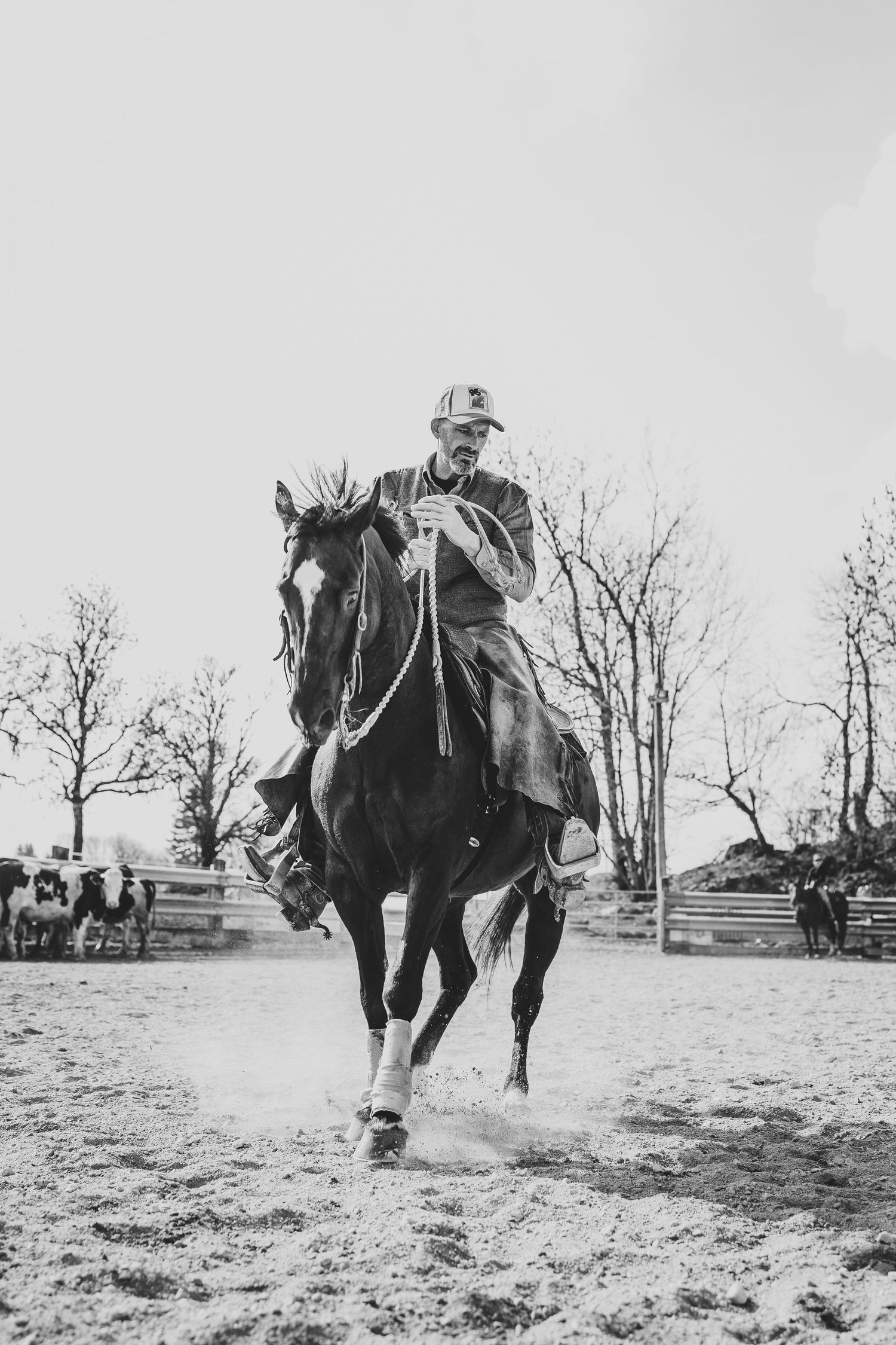 Black and white photo of a male rider galloping a dark horse in an outdoor arena.