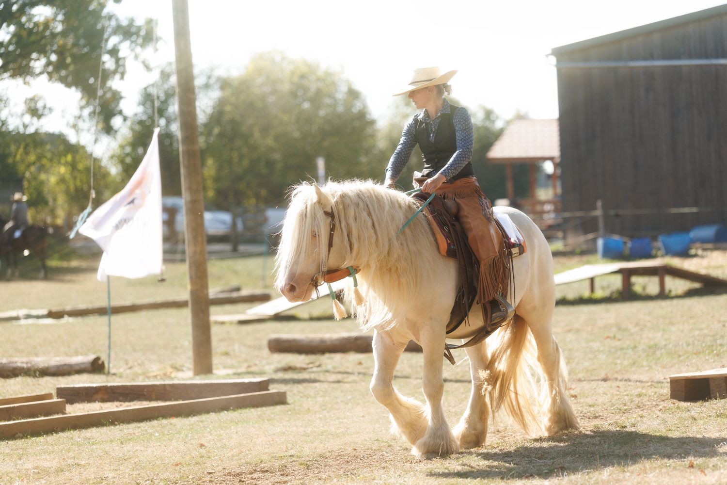 Person riding a pale palomino horse in a sunny outdoor arena wearing a cowboy hat during an equestrian event.