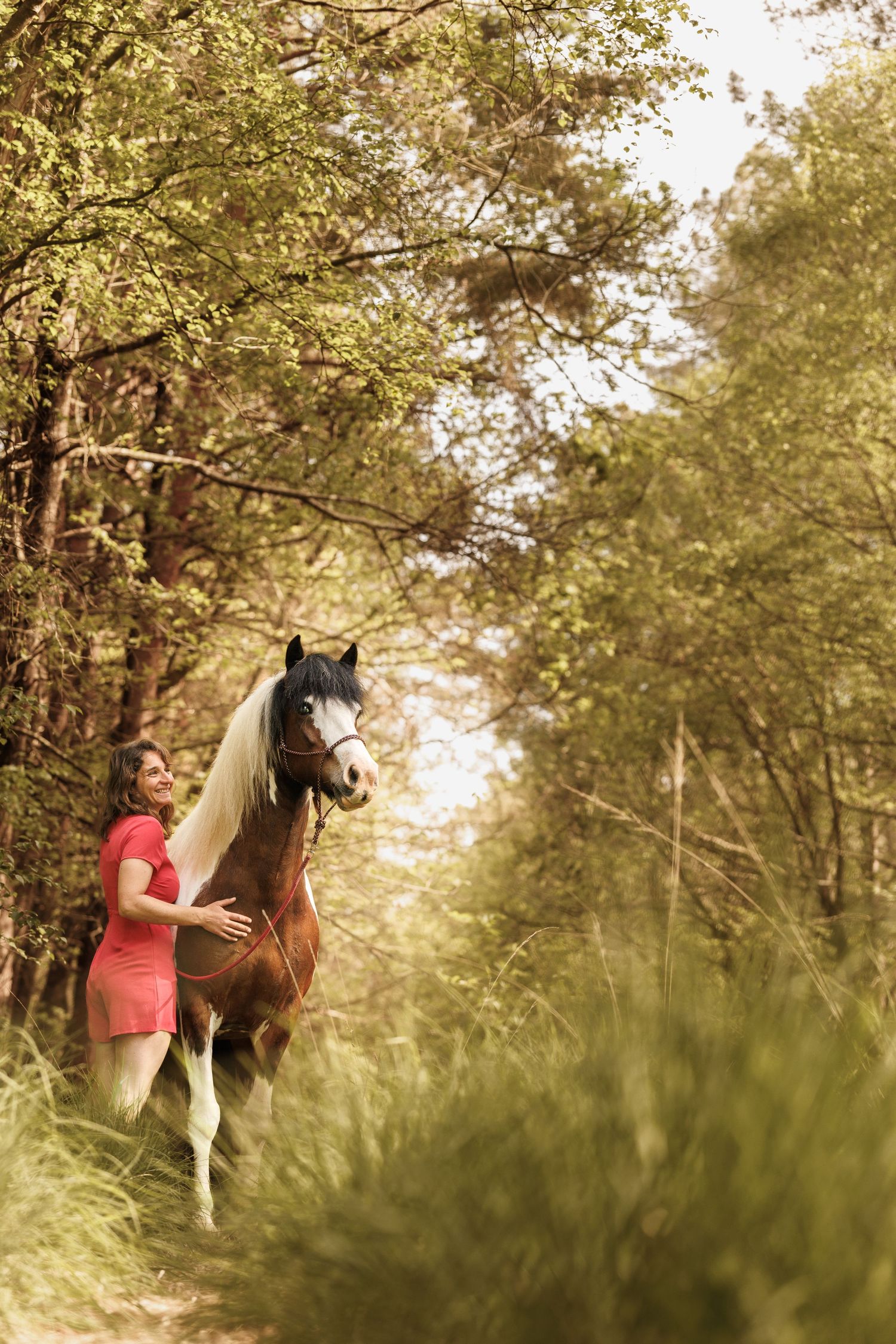 Woman in red dress walking with a brown and white horse along a forest path surrounded by lush green trees.