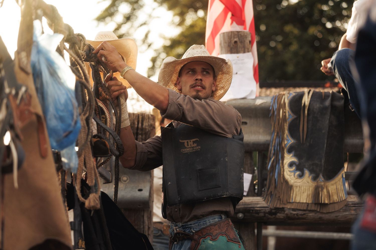 Cowboy in white hat and vest prepares bull riding equipment at outdoor rodeo event with flag in background.