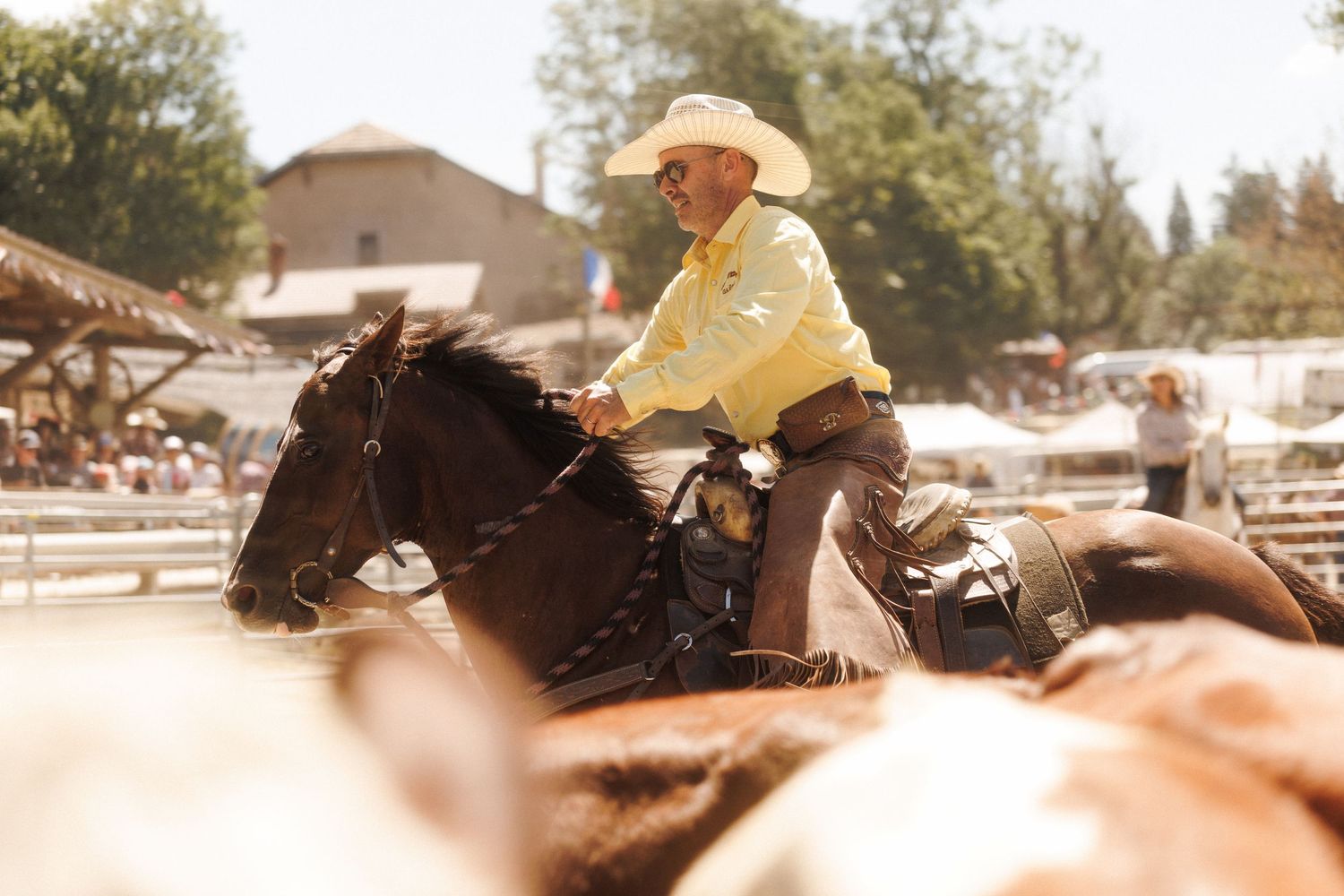 A cowboy in a yellow shirt and white hat rides a dark horse at a sunny outdoor rodeo event.