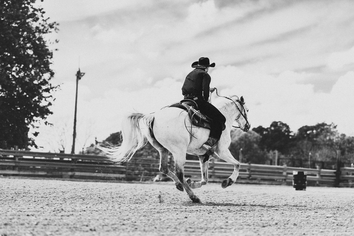 A cowboy in a black hat rides a white horse at full gallop in a sandy arena during an equestrian event.