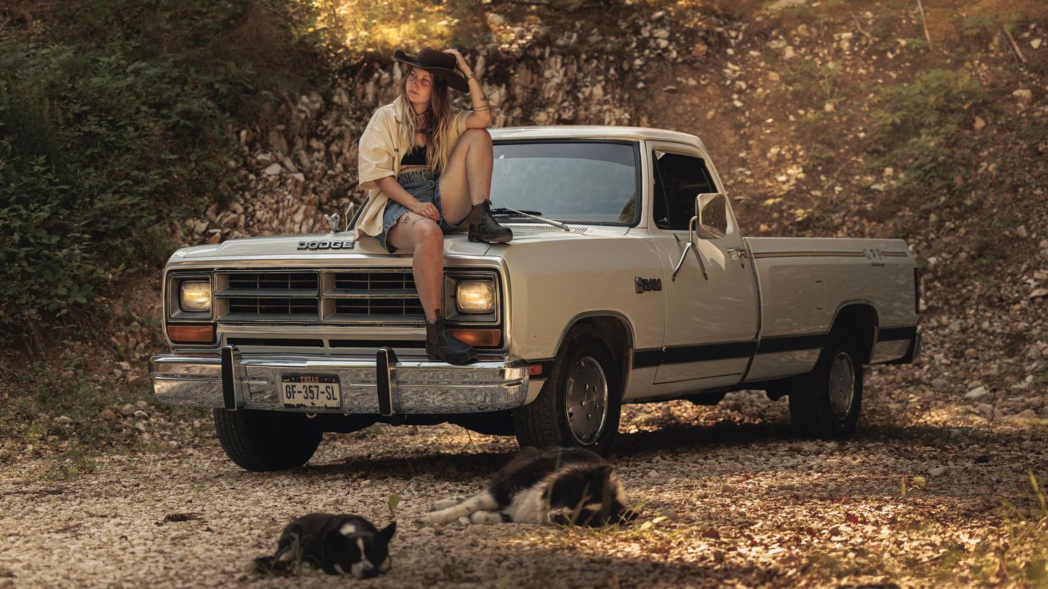 Two women sitting on the hood of a vintage white Dodge pickup truck outdoors, with cats resting on the ground nearby.
