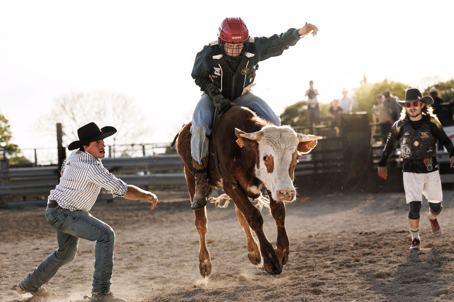 A rodeo rider in a red helmet bull riding while two cowboys assist during an outdoor rodeo event.