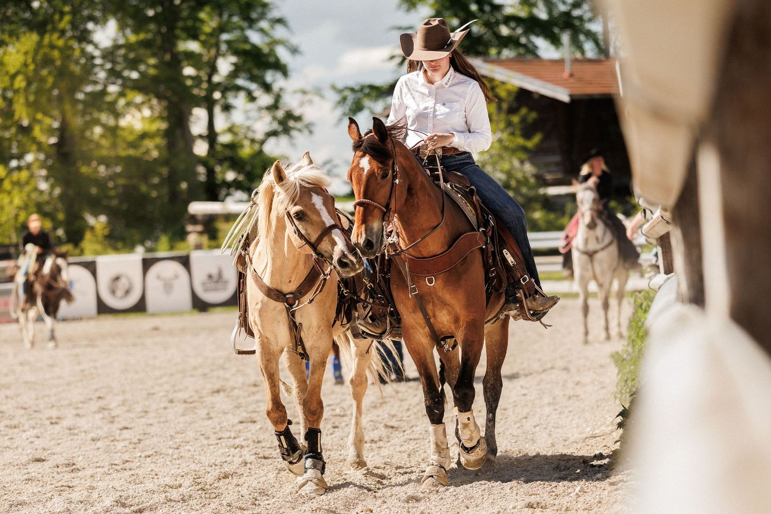 A rider in western attire guides two horses through an outdoor equestrian arena surrounded by trees.